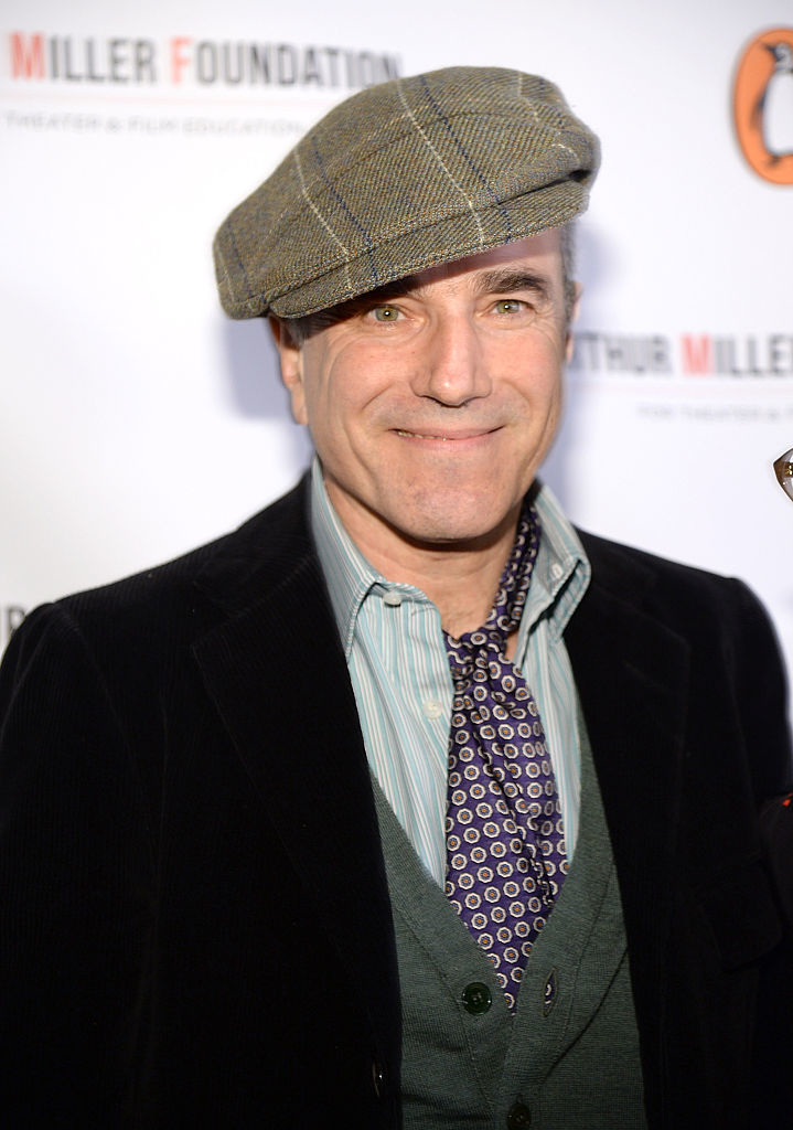 Daniel Day-Lewis at an event, wearing a plaid flat cap, a striped shirt, a black velvet jacket, and a patterned tie. The backdrop features "Miller Foundation" text