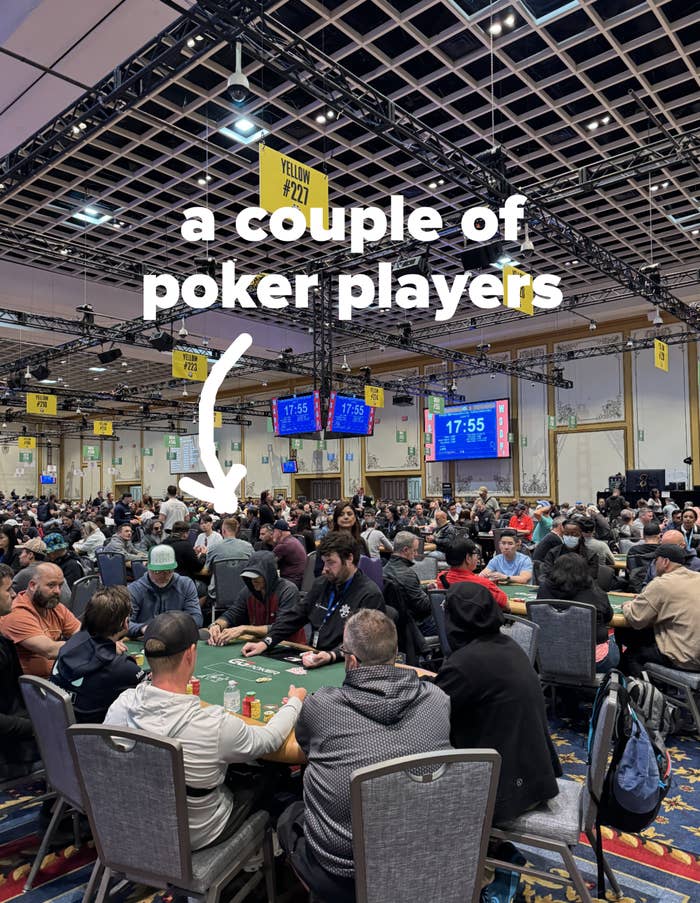 Large poker tournament in a casino with many players sitting at tables under hanging yellow signs indicating table numbers. Timer displays on screens