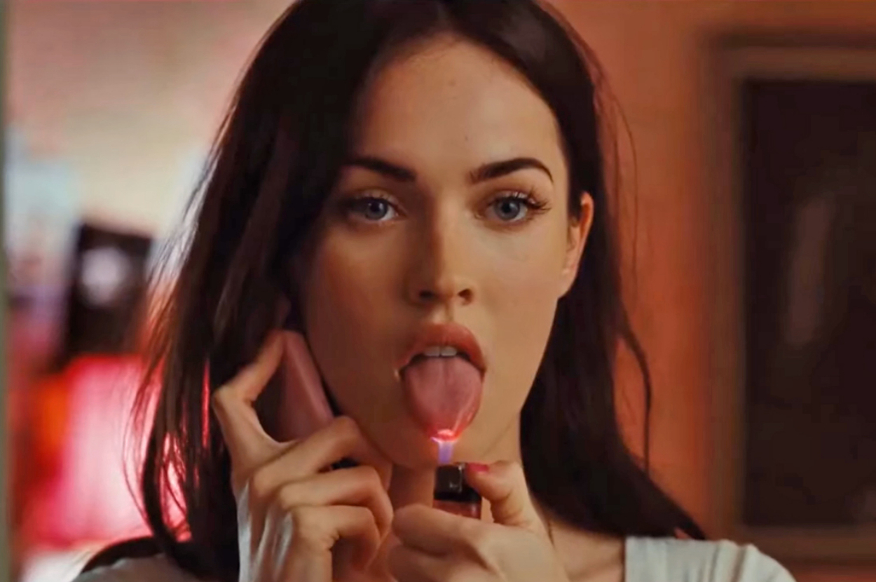 Megan Fox in a close-up shot, holding a phone with one hand and a lighter near her tongue with the other as Jennifer in Jennifer's Body