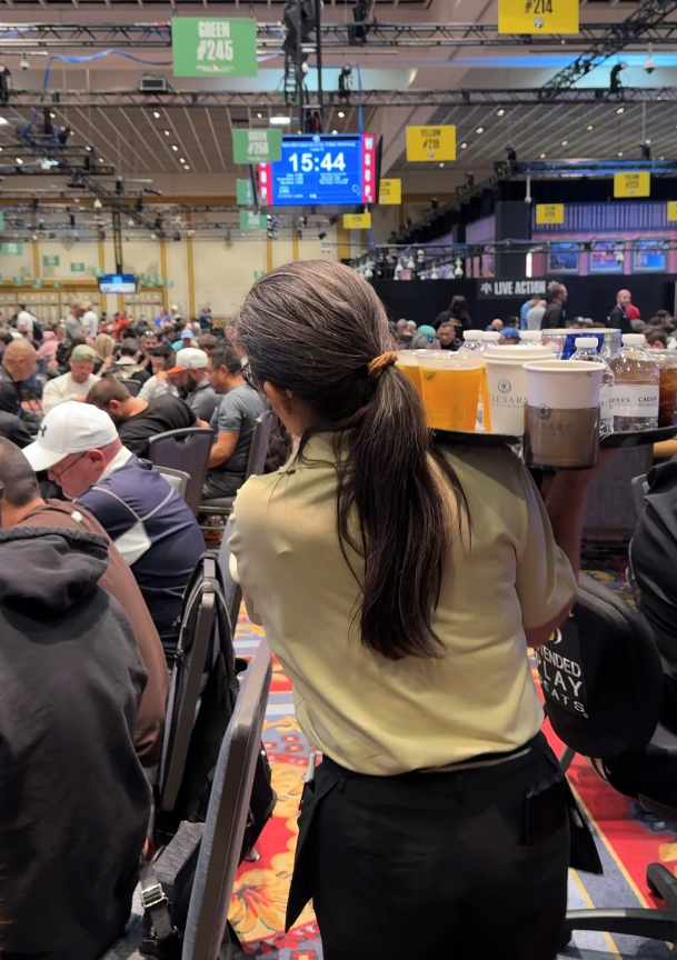 A server carrying a tray of beverages at a large, busy event with many attendees seated at tables. The server has long hair tied back and wears a uniform