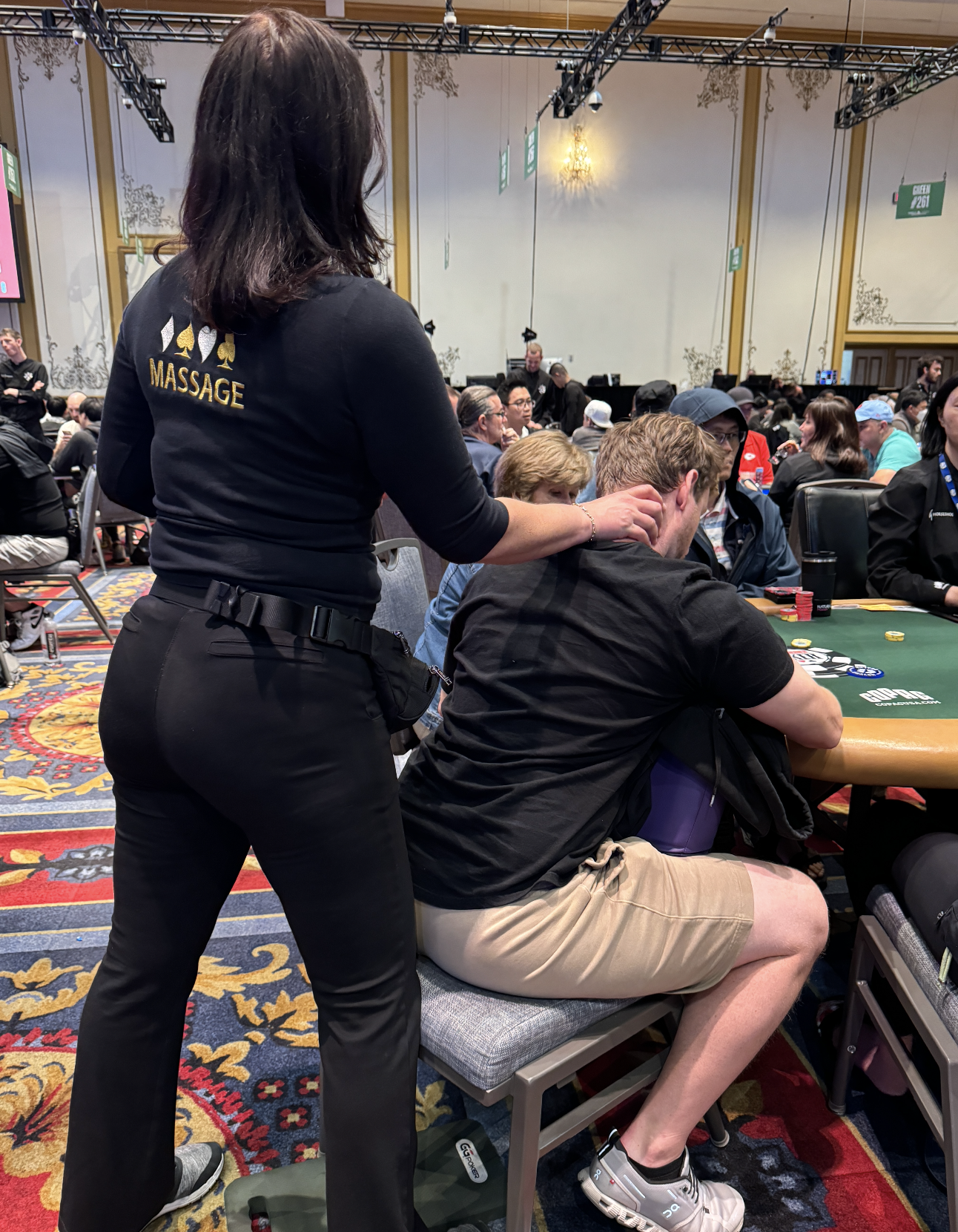 Two people at a poker table; a seated man in a black shirt, shorts, and a massage therapist standing behind him, massaging his neck, with "Massage" on her shirt