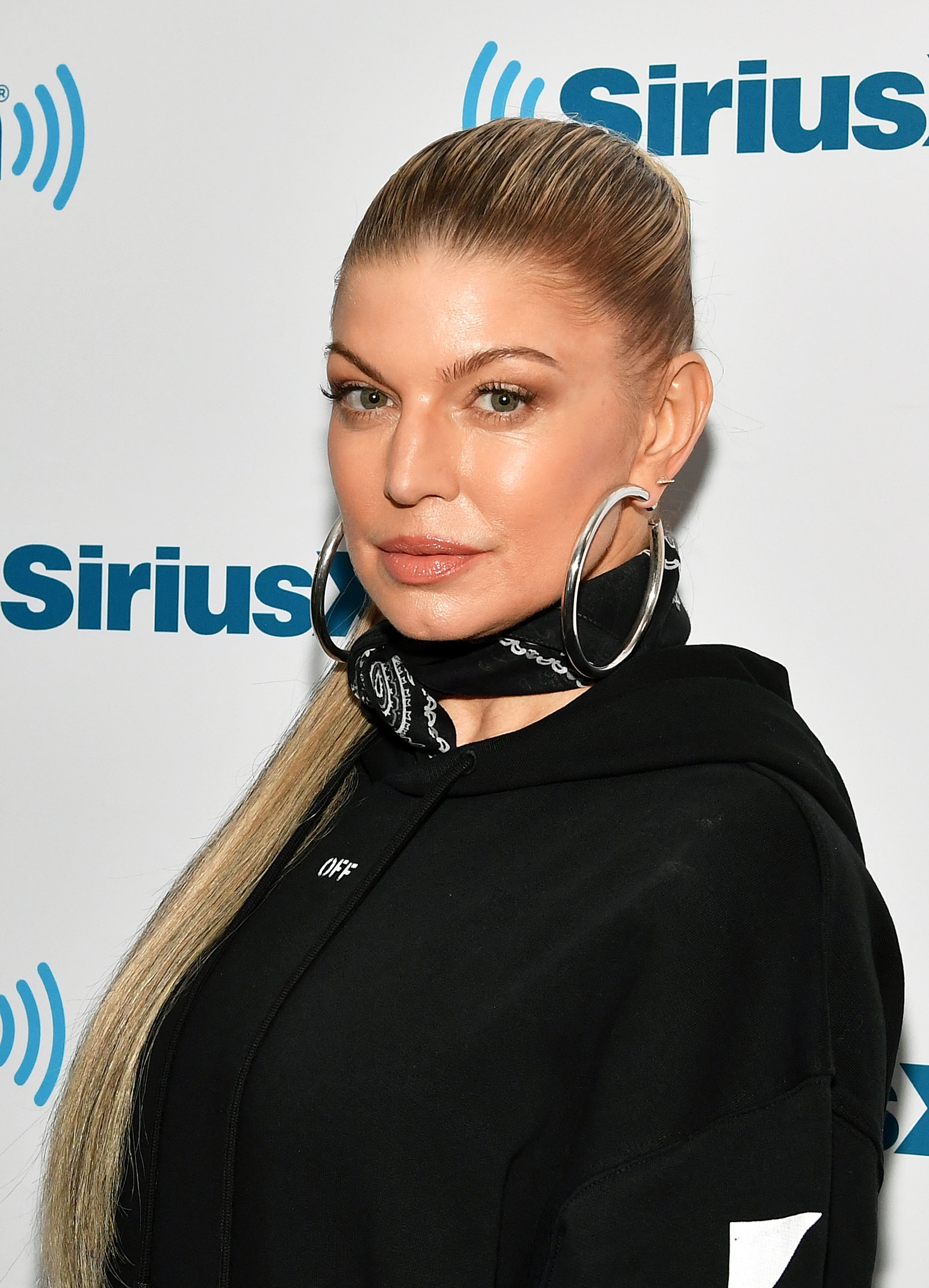 Fergie at a Sirius event wearing a sleek black hoodie, large hoop earrings, and a bandana around her neck, with her hair pulled back in a high ponytail