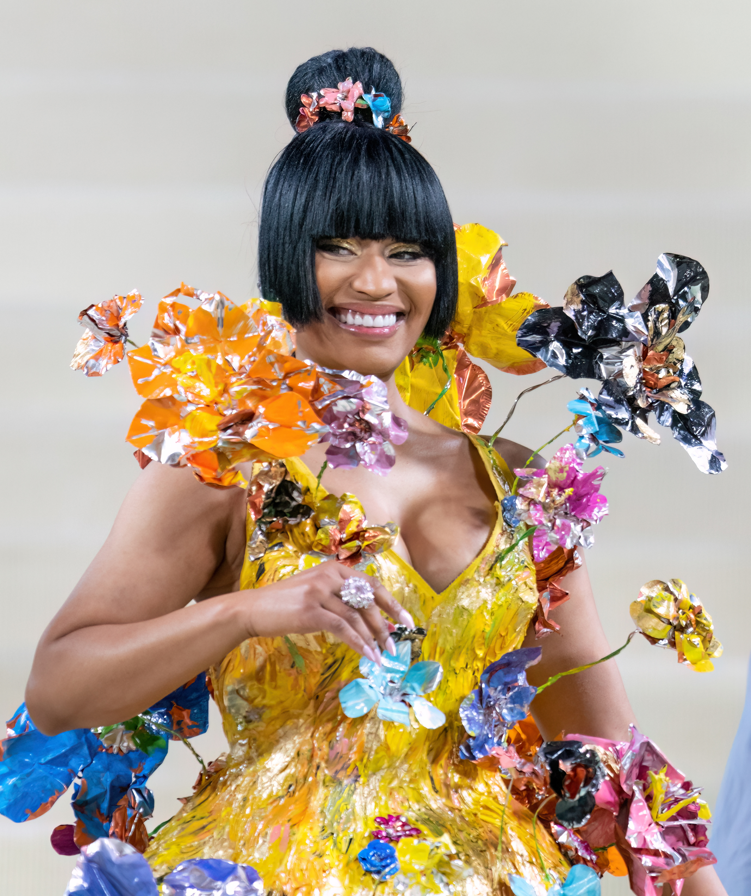 Nicki Minaj smiling, wearing a vibrant dress adorned with large, colorful, flower-like decorations and a high bun hairstyle with bangs