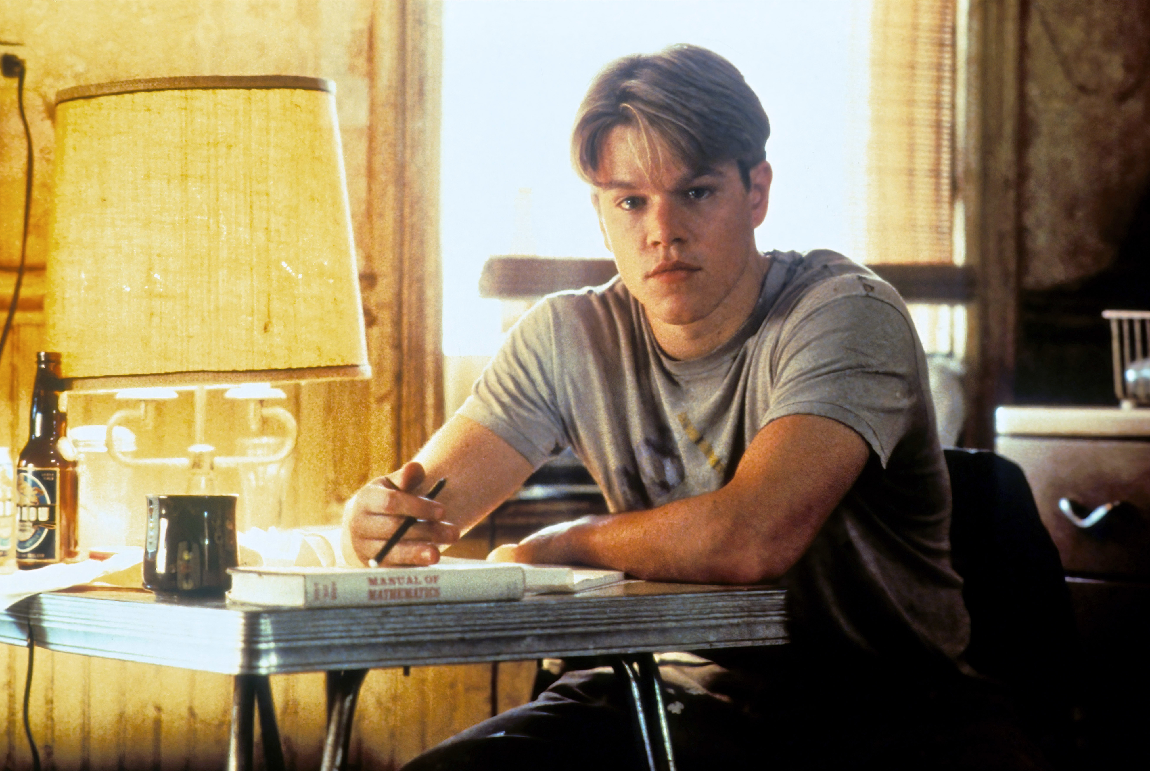 Matt Damon, in a casual t-shirt, sits at a table with a study book titled "Manual of Mathematics", a pen in hand, looking towards the camera