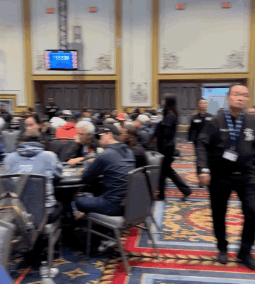 People are sitting at tables, participating in an event in a large, ornate room. There's a person standing and holding a microphone and a security person walking by