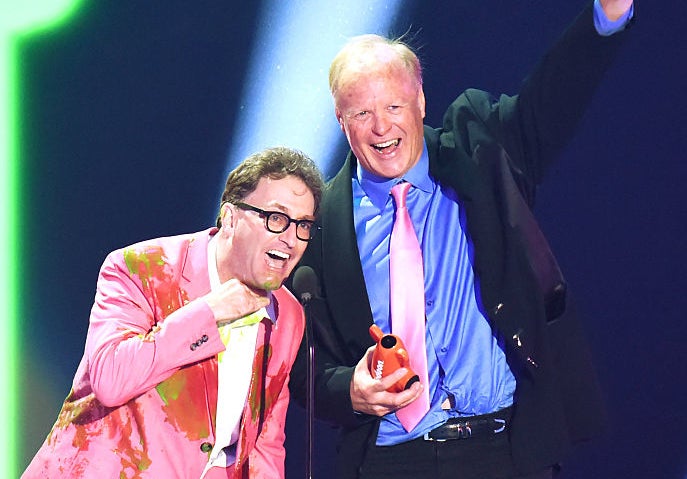 Stephen Colbert and Ed Begley Jr. on stage, with Ed wearing a suit and pink tie and Stephen in a colorful suit, sharing a moment at a celebrity event