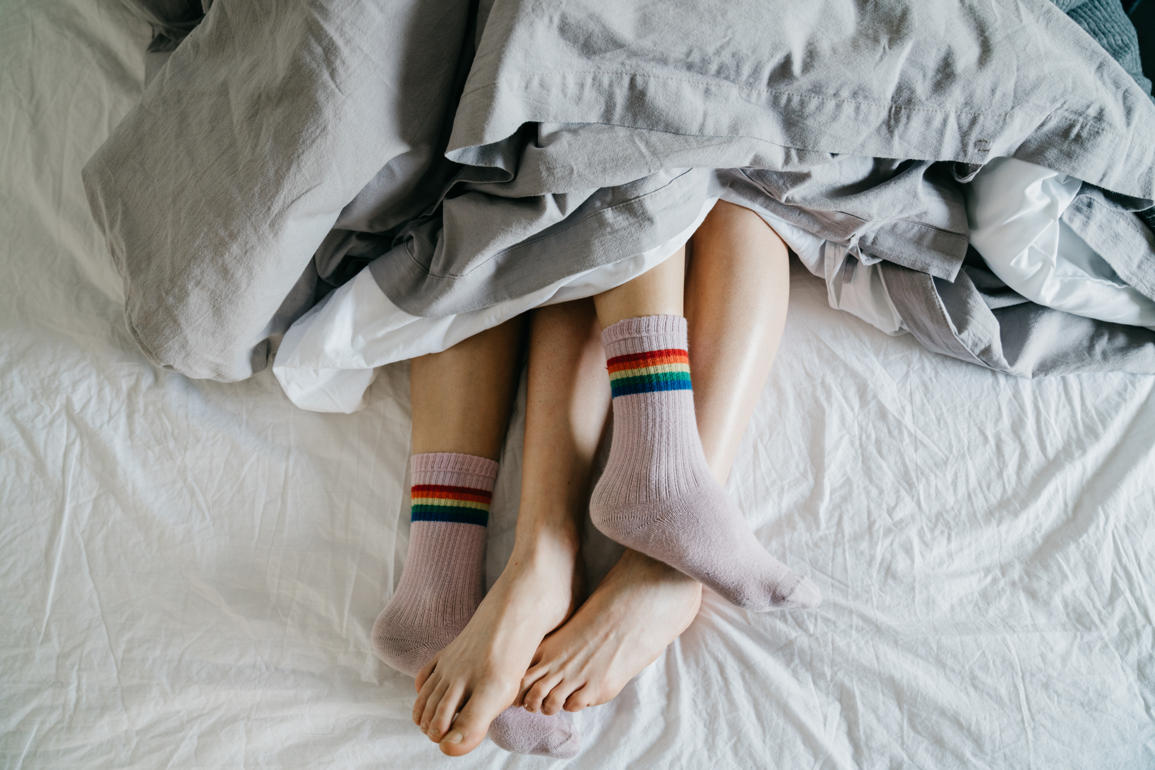 Two pairs of feet, wearing pastel socks with rainbow stripes and bare, peek from under a bedcover, suggesting a cozy scene. No names or text present