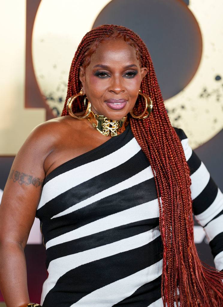 Mary J. Blige poses on a red carpet, wearing an off-the-shoulder striped dress, gold choker, and large hoop earrings