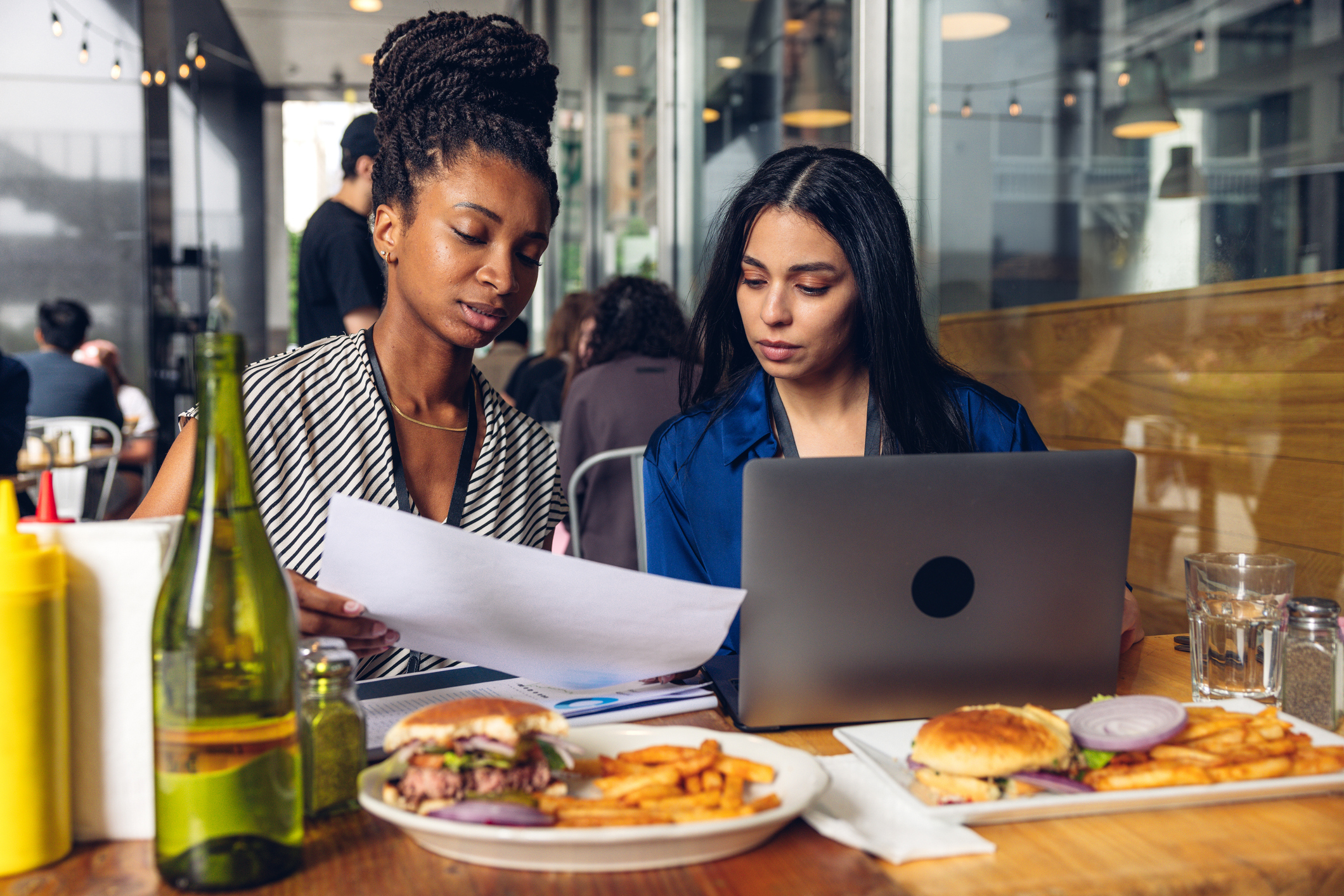 Two women reviewing documents at an outdoor café table, with burgers and fries in front of them, a laptop open, and condiments and a green bottle nearby