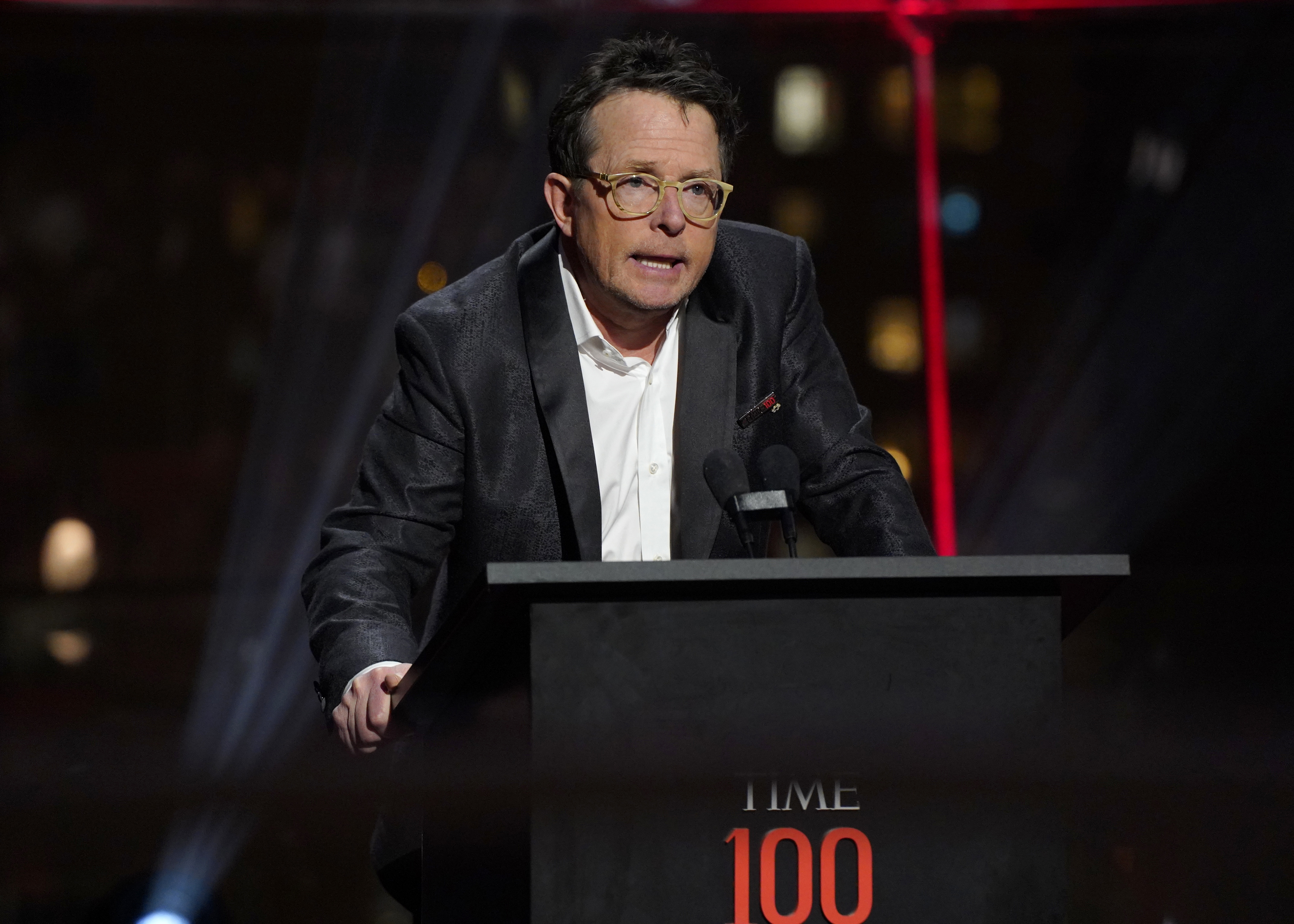Michael J. Fox speaking at a podium during the TIME 100 event, wearing a dark suit and glasses