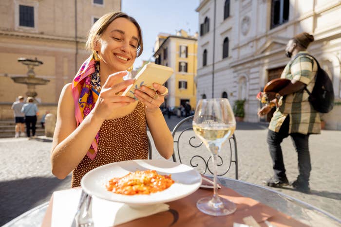Person smiling while looking at phone, with a plate of risotto and a glass of white wine at an outdoor café in a European plaza