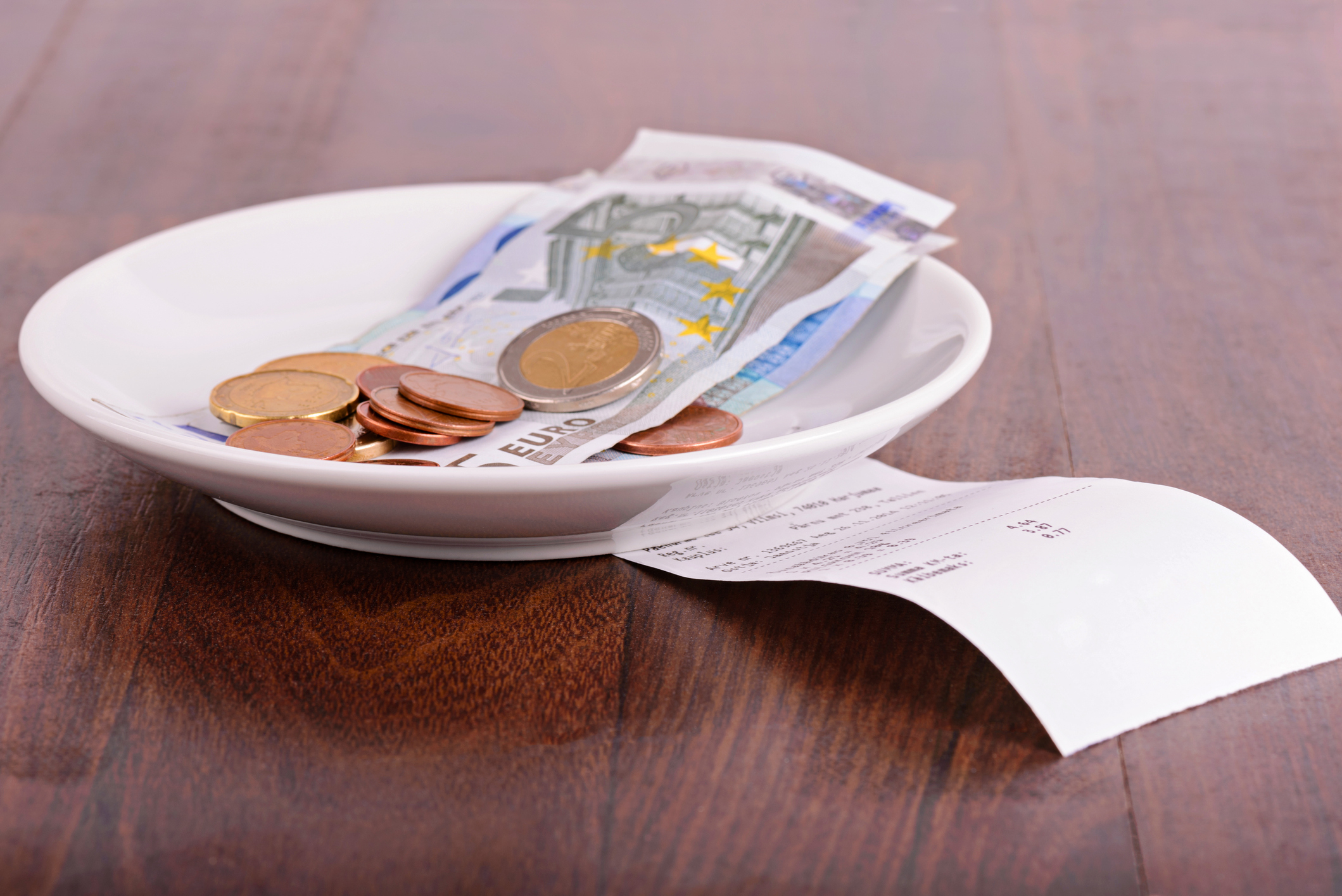 A white dish with various euro banknotes and coins on a wooden table alongside a receipt