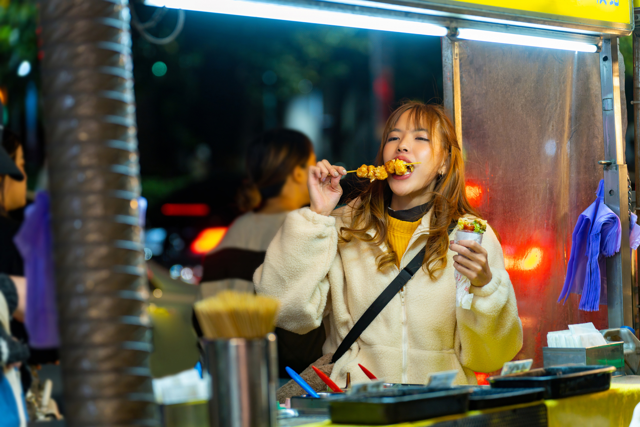 A person enjoys street food at a night market, biting into a skewer and holding a cup with other snacks. Various food items and utensils are on the counter
