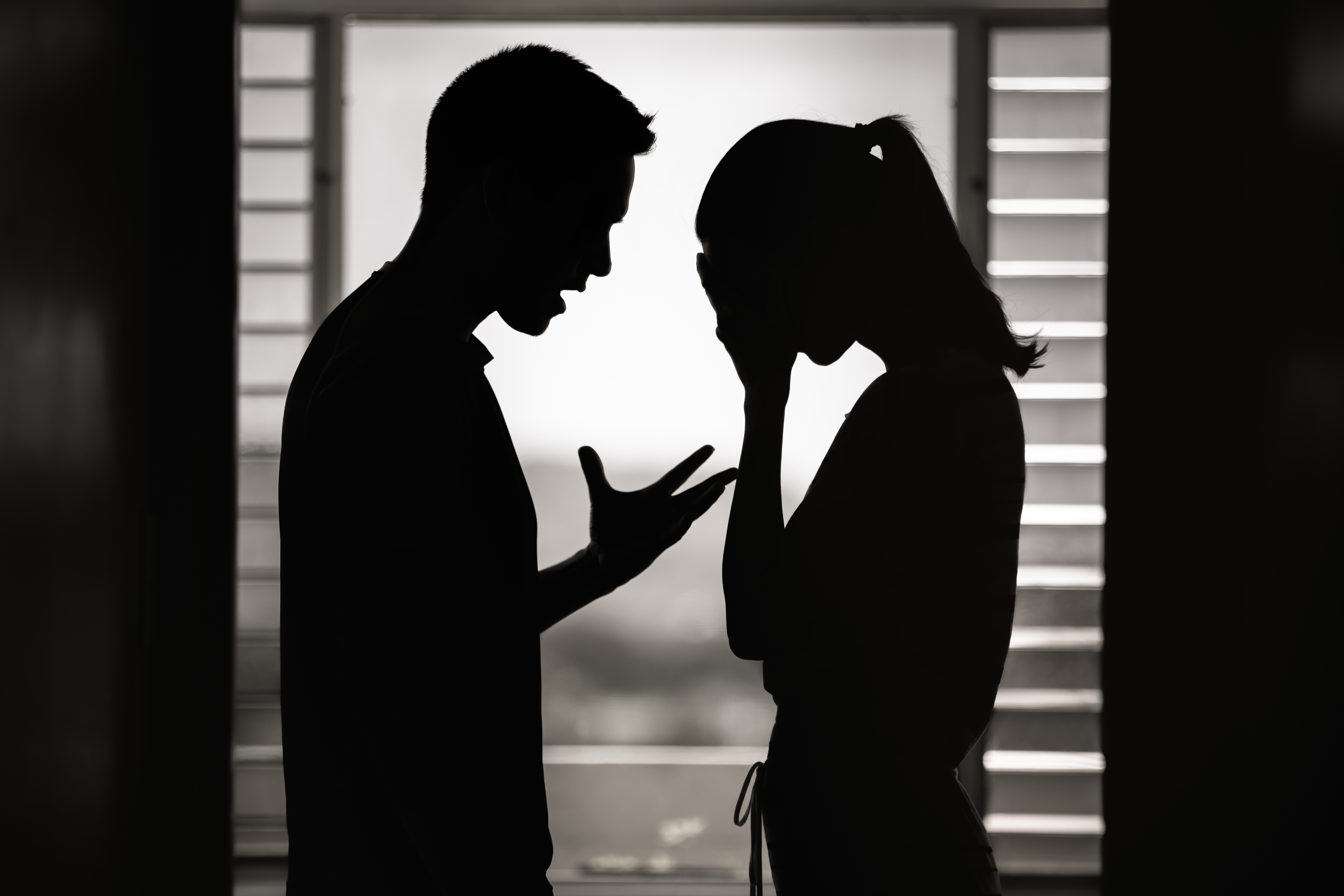 Two people in silhouette face each other. The person on the left is gesturing angrily, while the person on the right covers their face with their hands