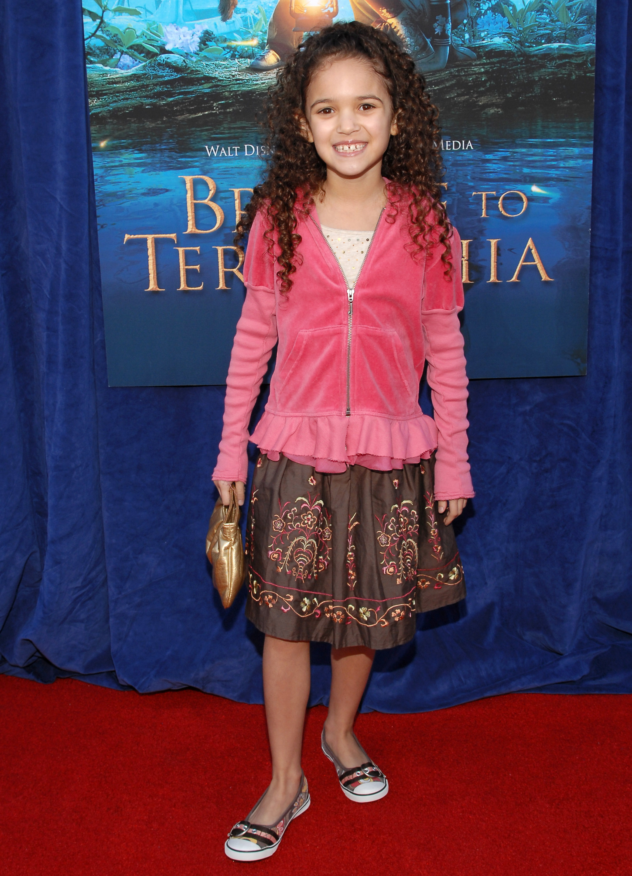 Madison Pettis on the red carpet at the "Bridge to Terabithia" premiere, wearing a pink jacket over a patterned skirt, holding a gold clutch