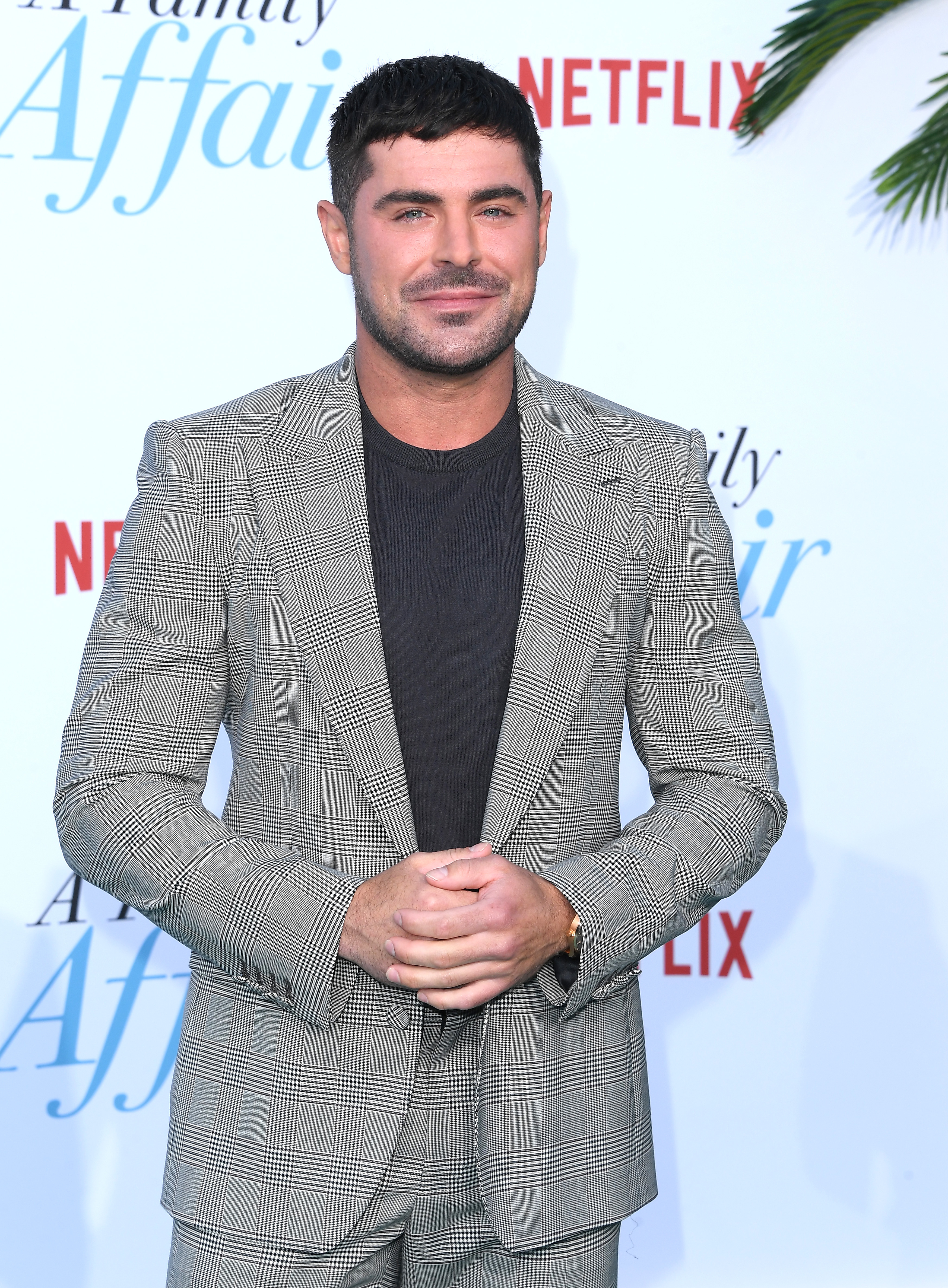 Zac Efron on the red carpet, wearing a gray checkered suit with a black shirt, at a Netflix event for "A Family Affair"
