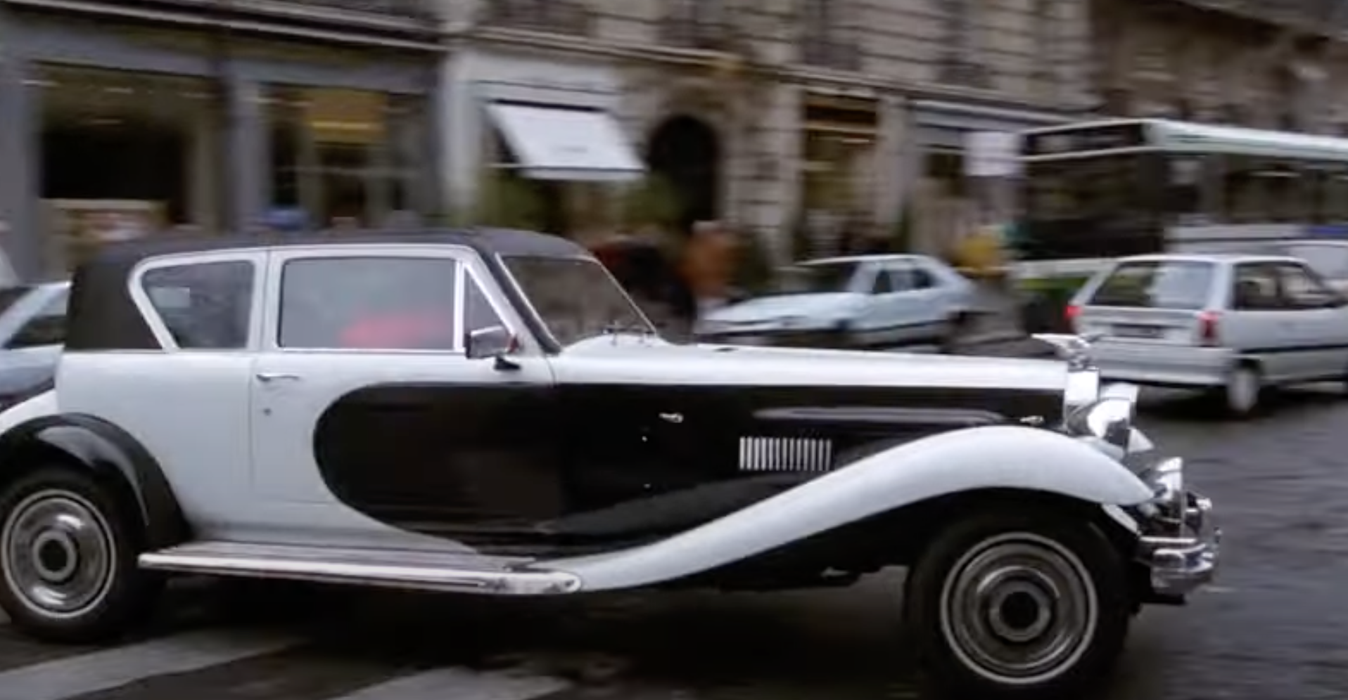 A classic black and white luxury car is driving down a busy city street with other cars and buildings in the background