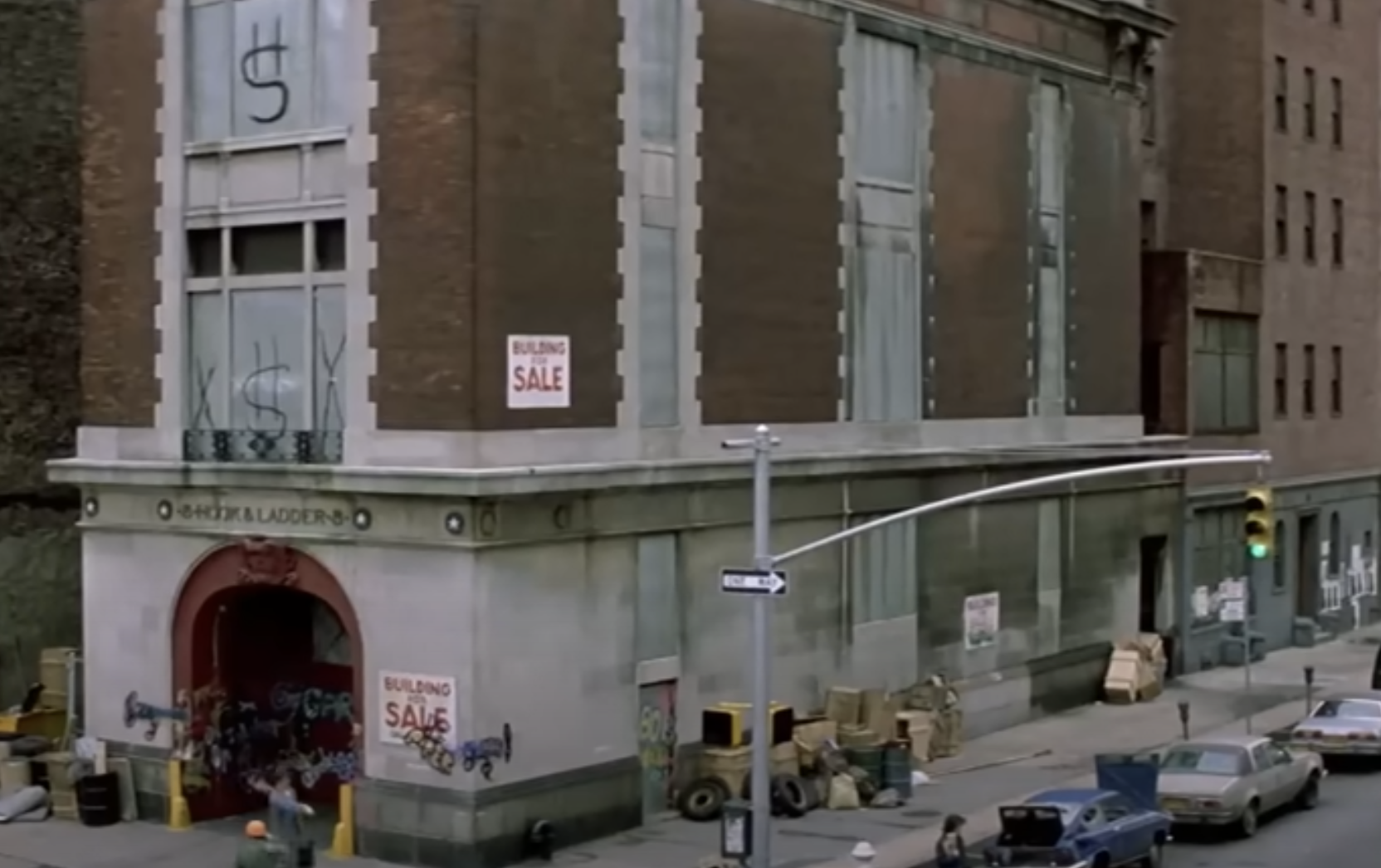 An old firehouse with "Hook & Ladder" sign, boarded windows, and "Building for Sale" signs on both sides. Various objects and debris are outside