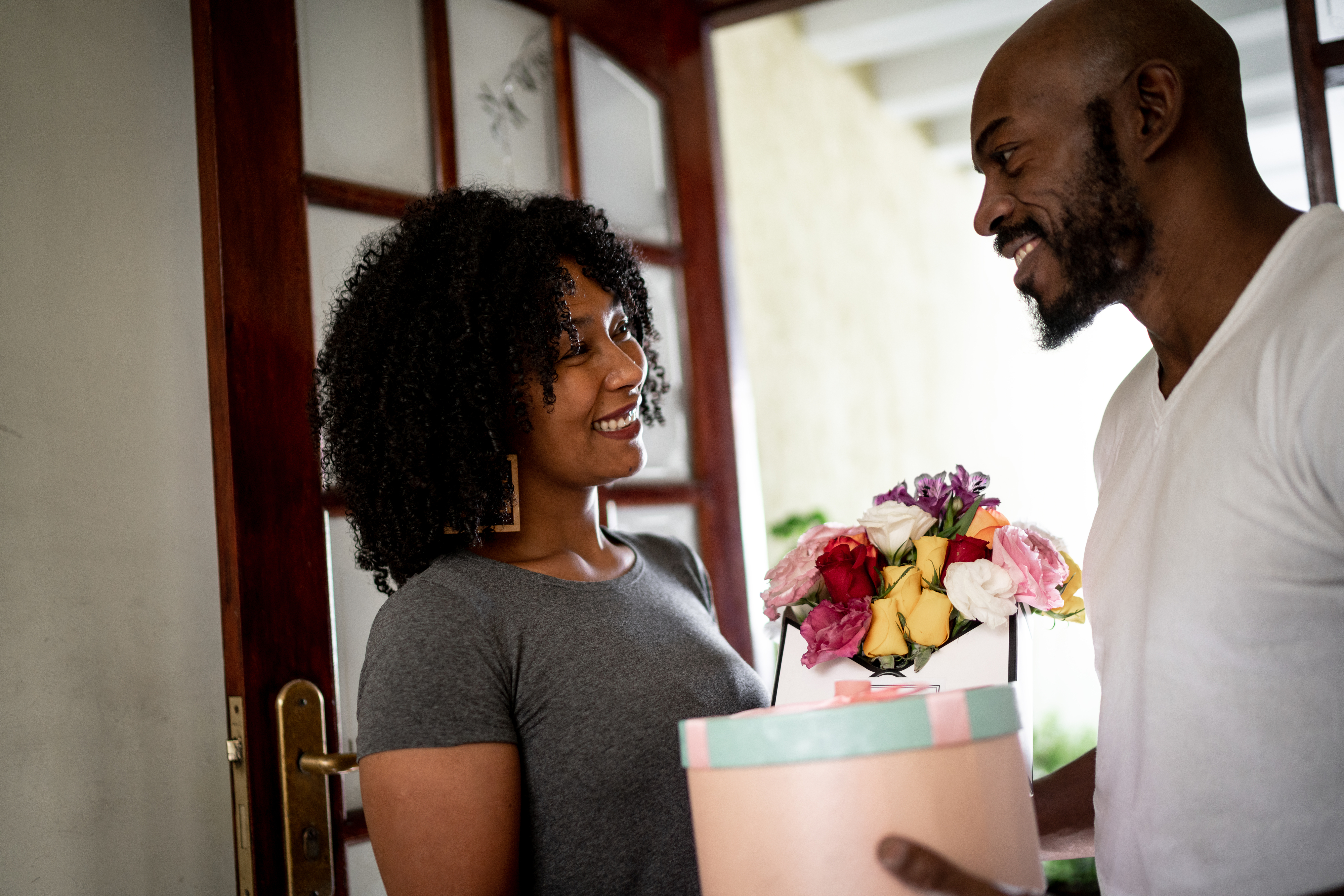Two people, a woman and a man, smiling at each other; she holds a bouquet of flowers, and he holds a gift box