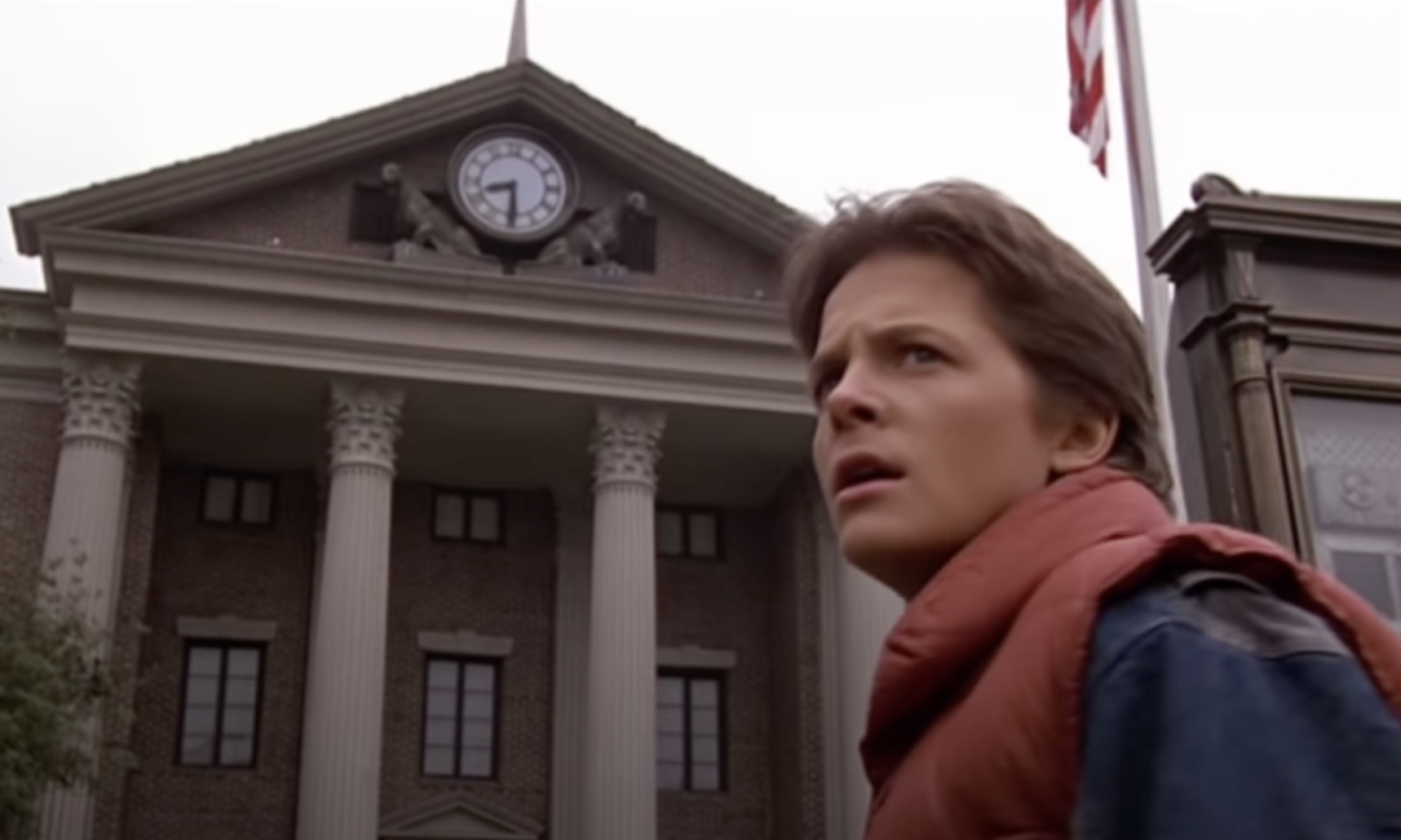 Michael J. Fox, wearing a puffy vest, stands in front of a courthouse with a clock tower