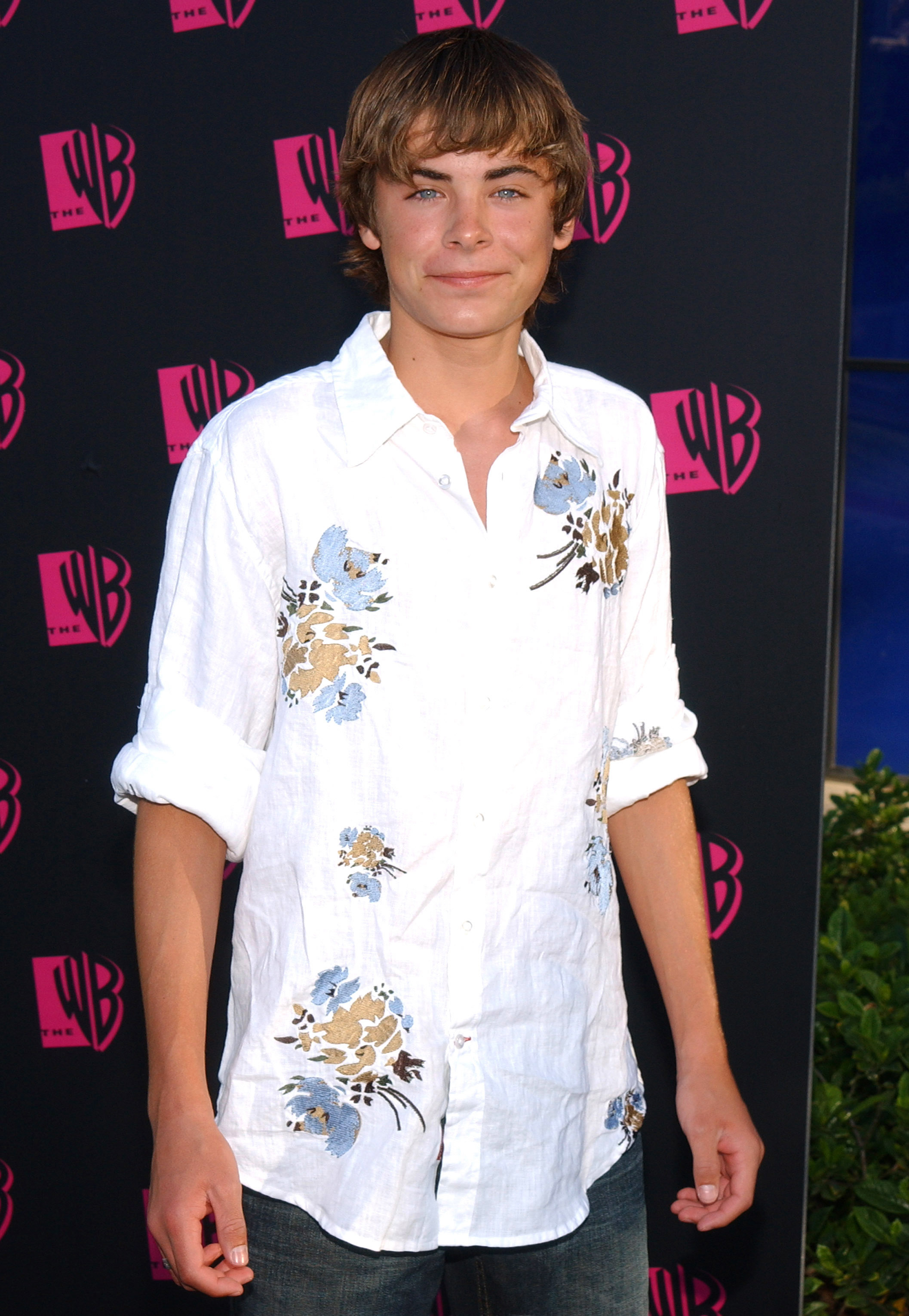 Zac Efron posing at a red carpet event, wearing a casual long-sleeved shirt with floral patterns and dark jeans