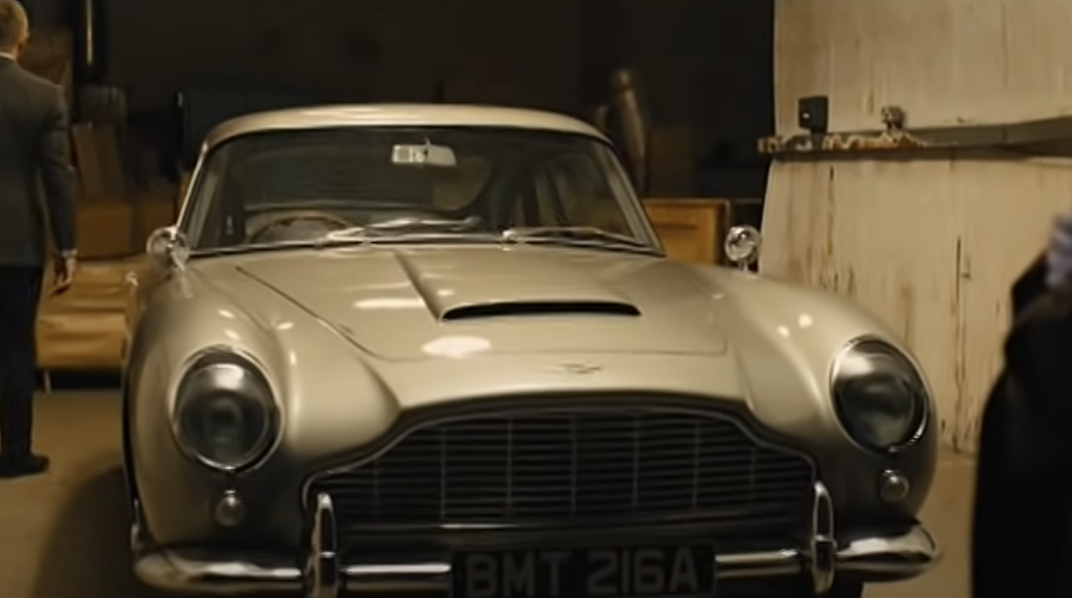 A classic Aston Martin car with license plate BMT 216A is parked in a garage. Two unidentified people are partially visible on each side of the frame