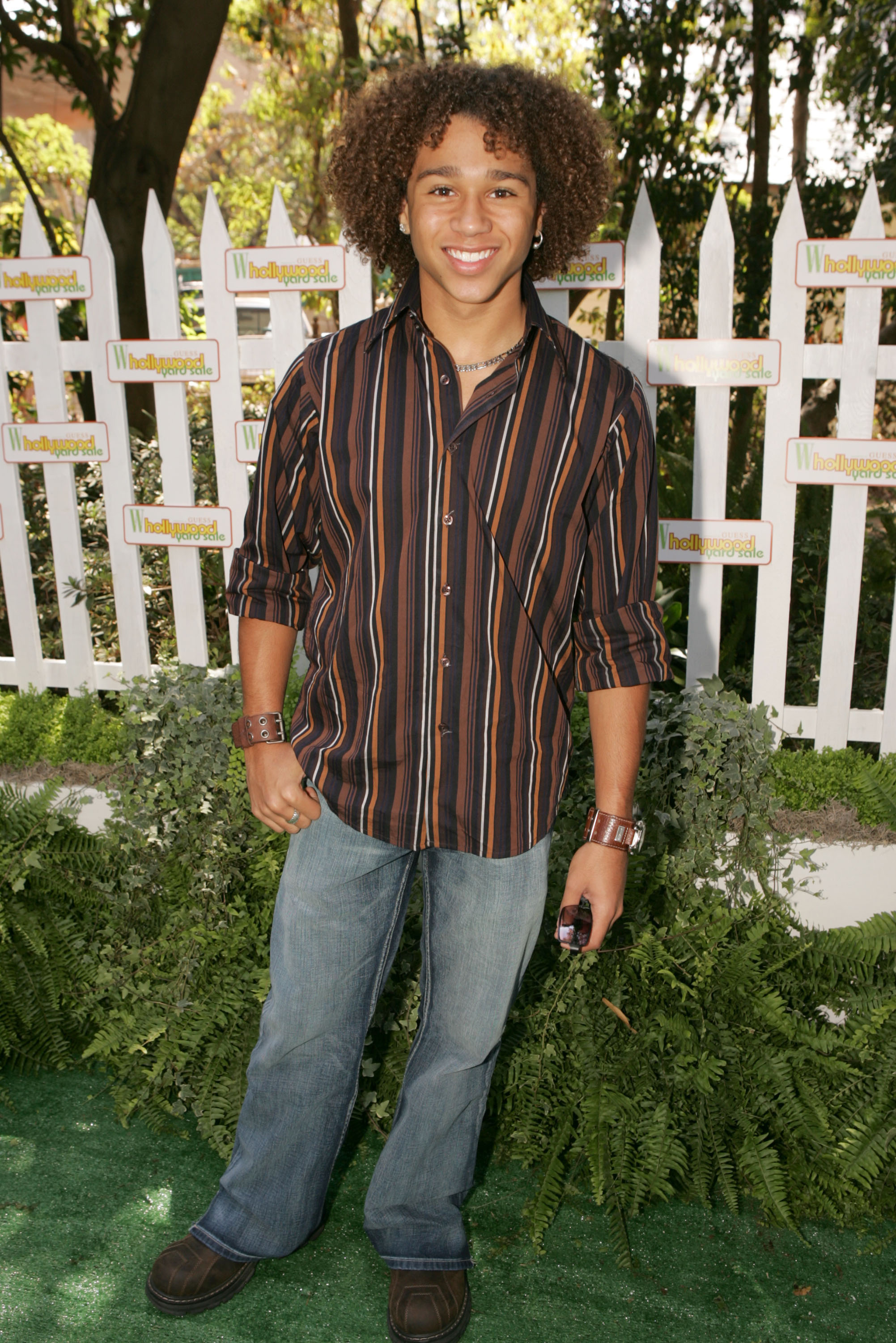 Corbin Bleu poses outdoors in a striped button-up shirt and jeans, smiling at the camera