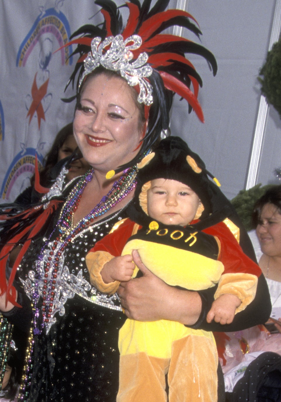 Camryn Manheim in an ornate outfit stands on a red carpet holding a baby in a Winnie the Pooh costume. Other children in costumes are in strollers nearby