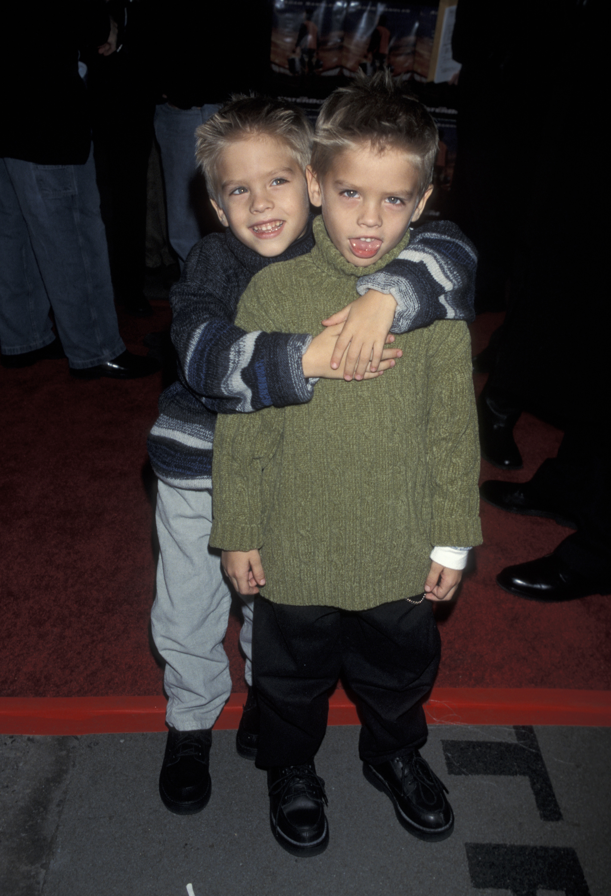 Dylan and Cole Sprouse are posing on a red carpet. Cole is standing behind Dylan, hugging him. They are both dressed in casual sweaters and pants