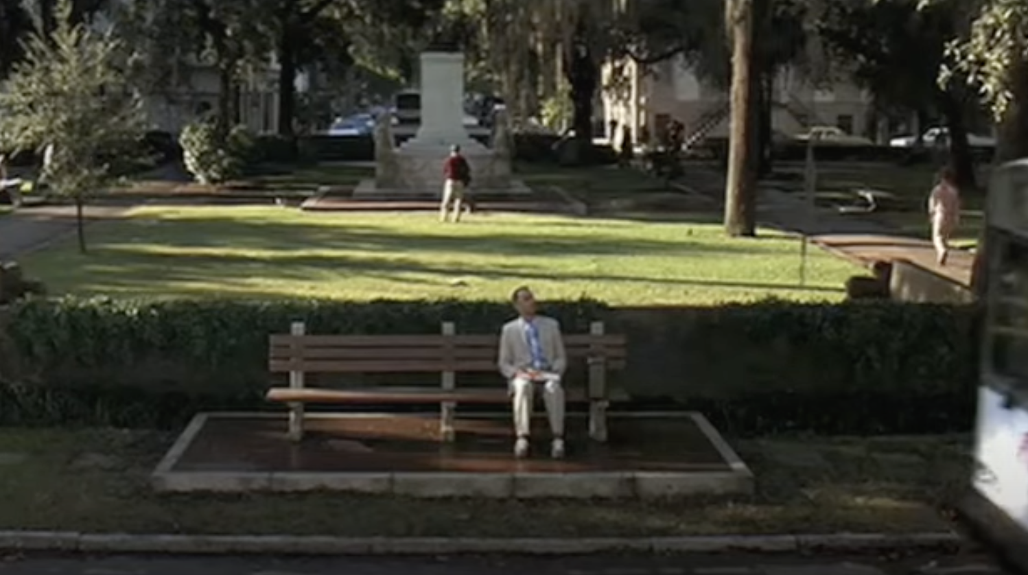 Tom Hanks, dressed in a light suit and tie, sits on a bench in a park with people walking and a white monument in the background