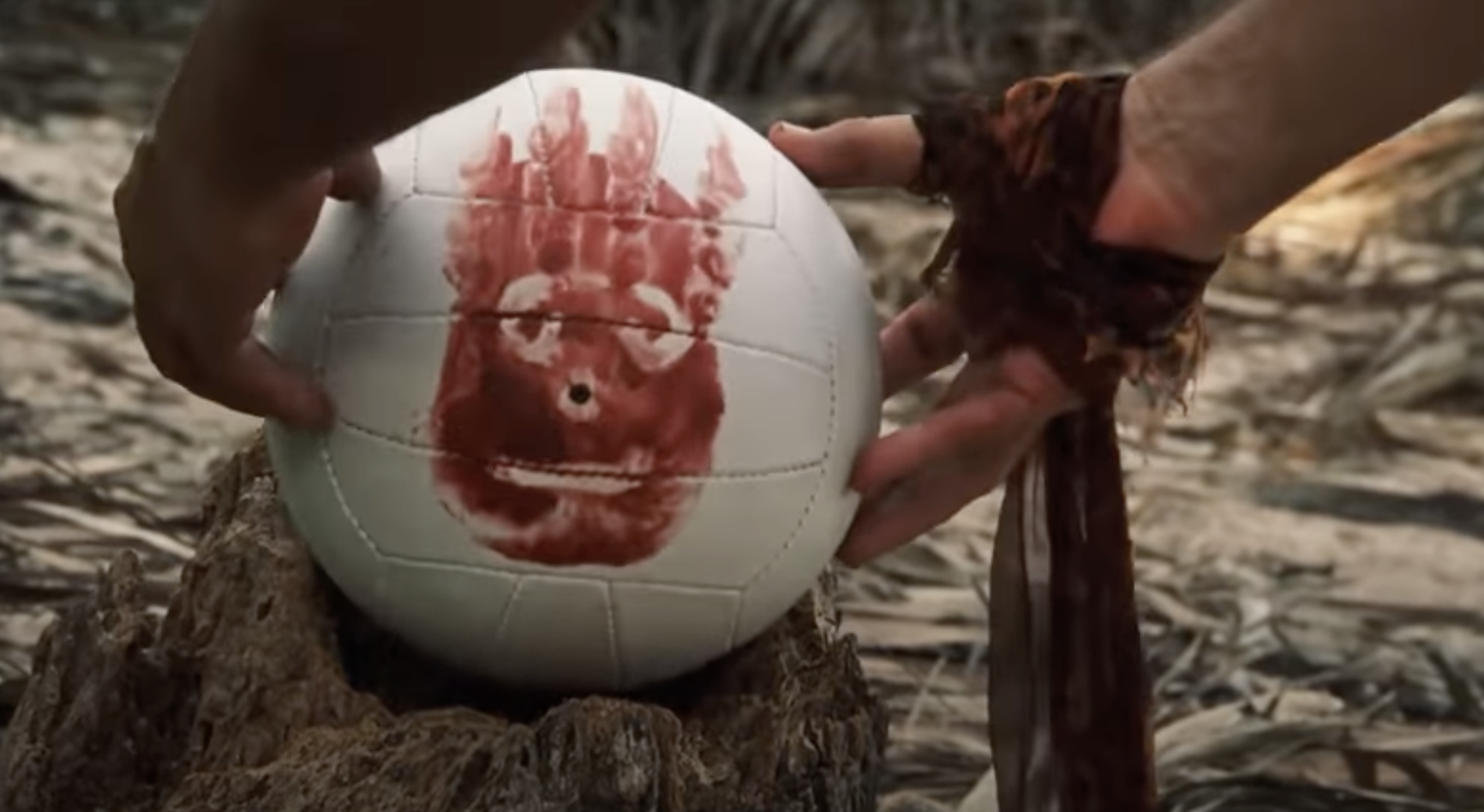 A person's hands place a volleyball with a red handprint face on a tree stump. The scene is from the movie "Cast Away," featuring the character Wilson