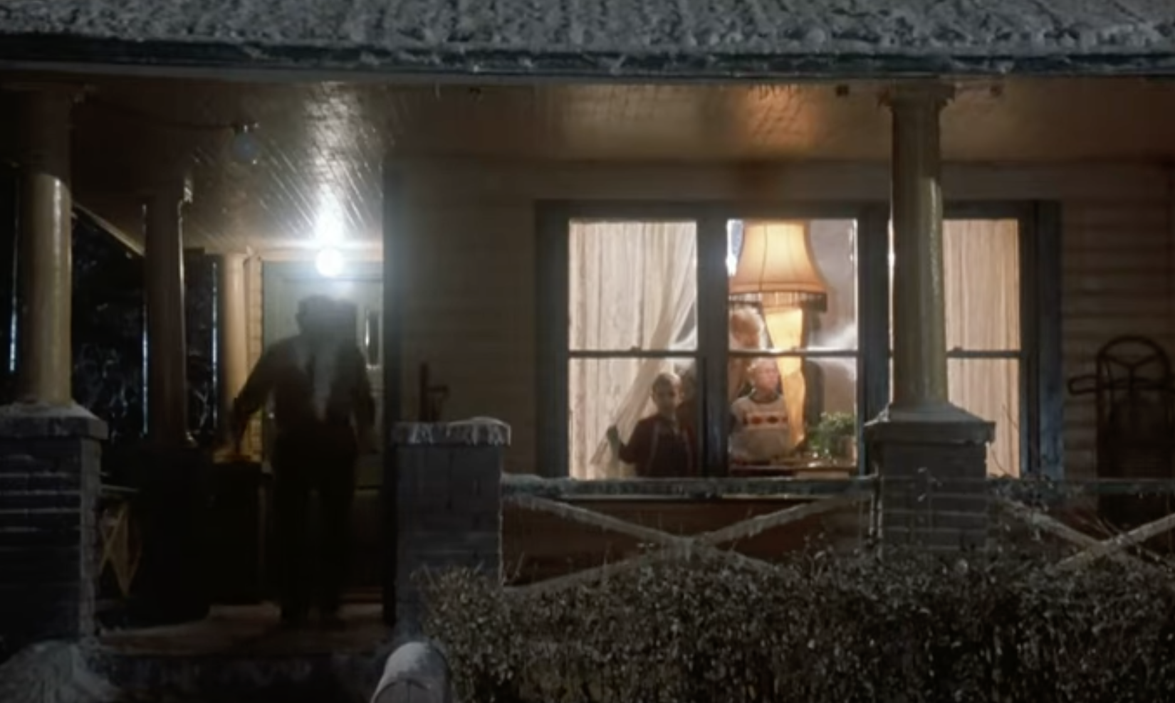 A scene from "A Christmas Story" showing a house with snowy surroundings, a man walking on the porch, and a family observing a leg lamp through the window
