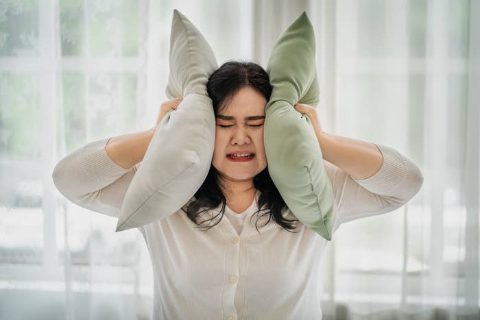 A woman in a white cardigan holds two pillows to her ears, looking frustrated. Background shows sheer curtains and a bright room