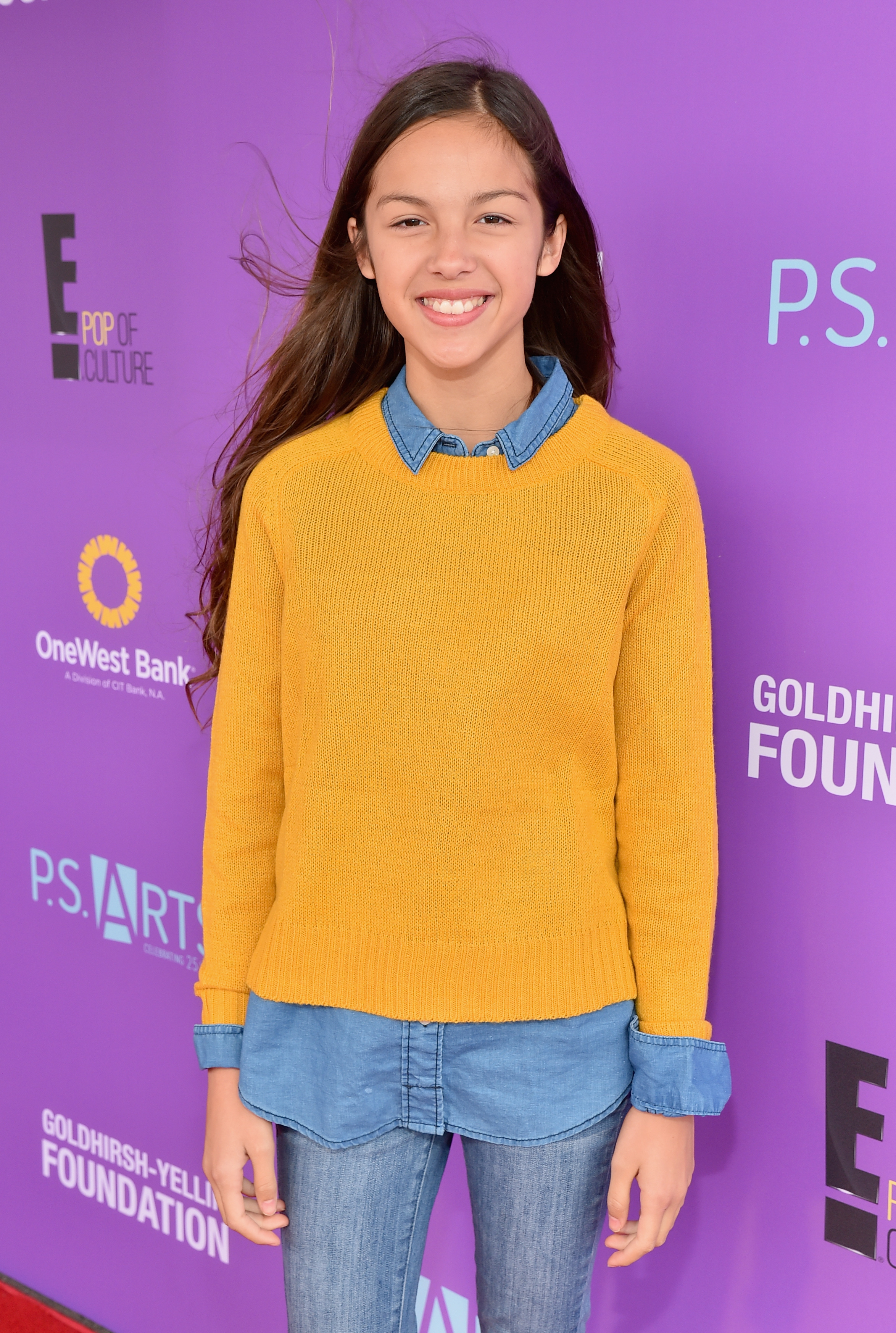 Olivia Rodrigo on the red carpet, wearing a yellow sweater over a denim shirt and jeans, posing in front of a backdrop for the Goldhirsh-Yellin Foundation event