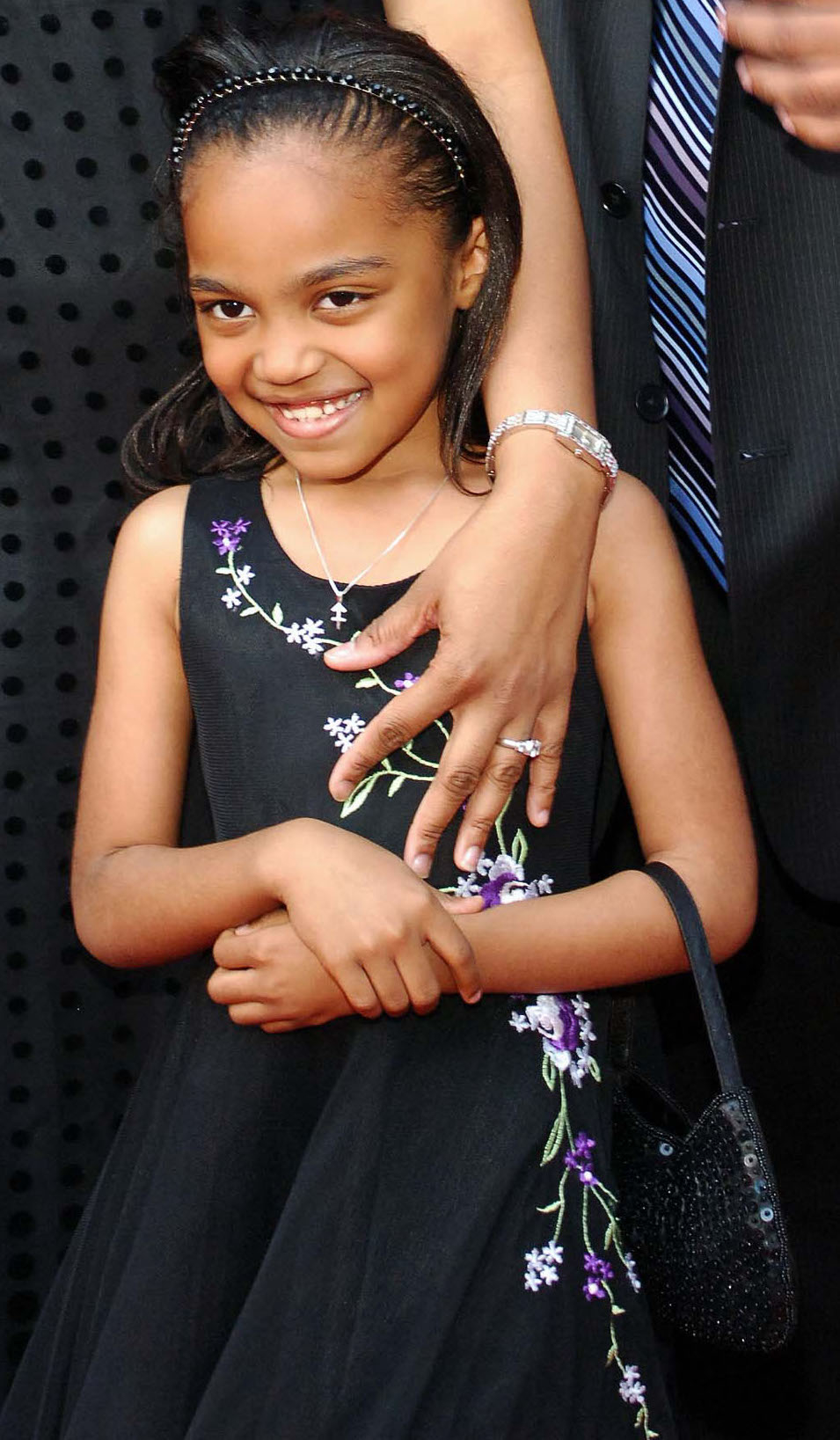 Young girl in a floral-patterned dress, smiling at the camera, holding a small black purse. An adult's arm, with a wristwatch and rings, rests on her shoulder