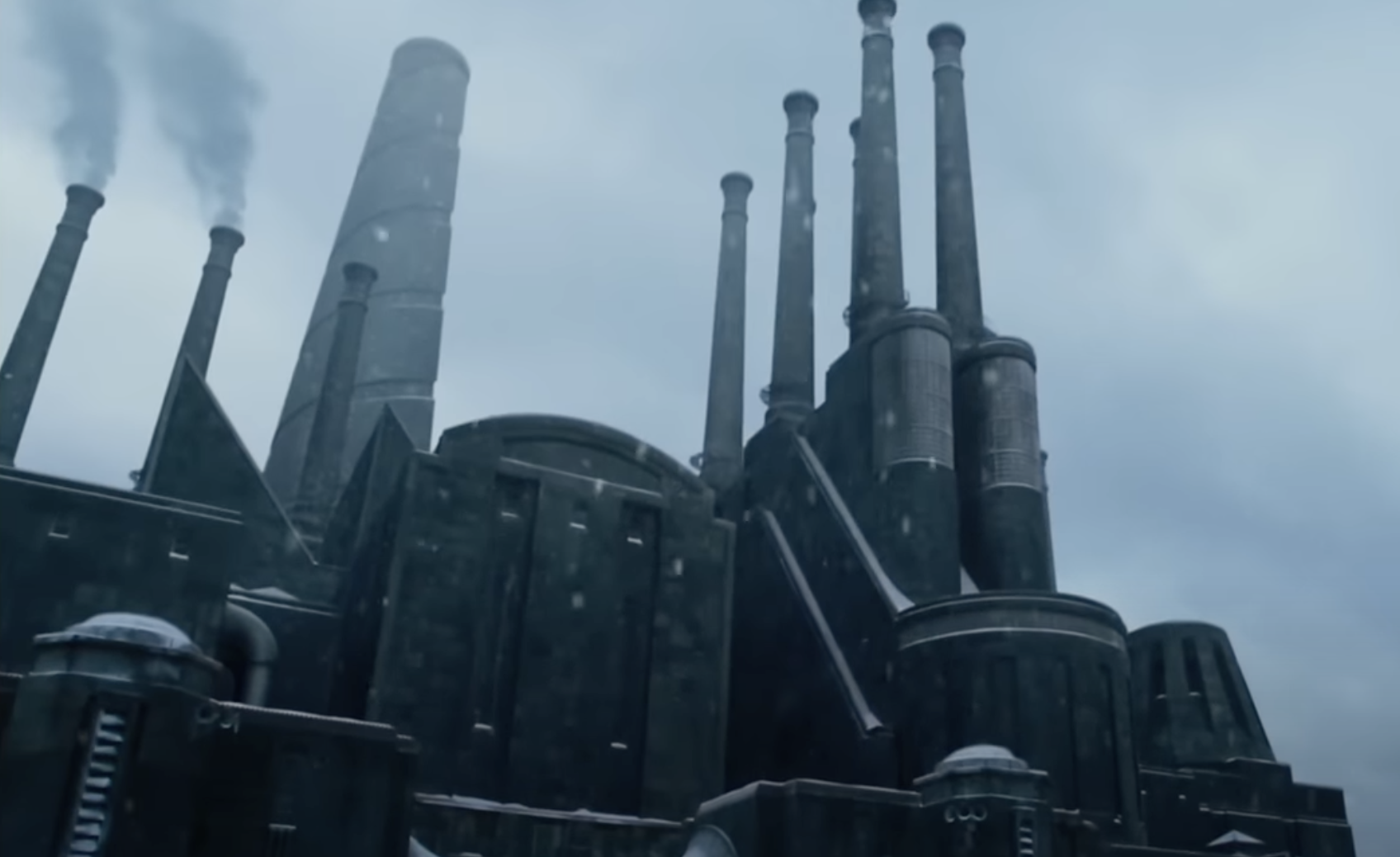 A large, industrial factory complex with numerous towering smokestacks emitting smoke under a cloudy sky