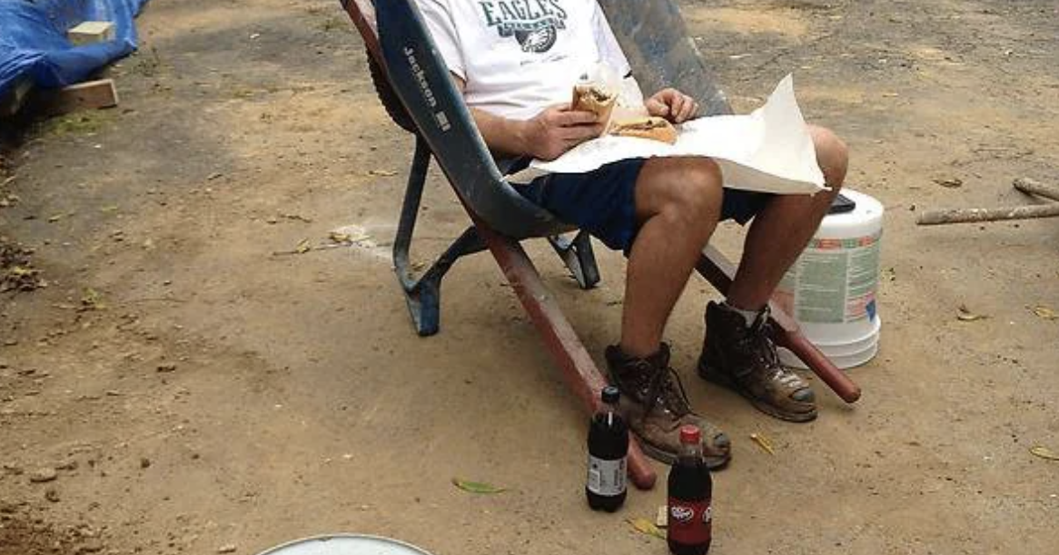 A person is sitting on a makeshift chair outdoors, eating a sandwich. Two soda bottles are on the ground near them. The shirt reads "Eagles"