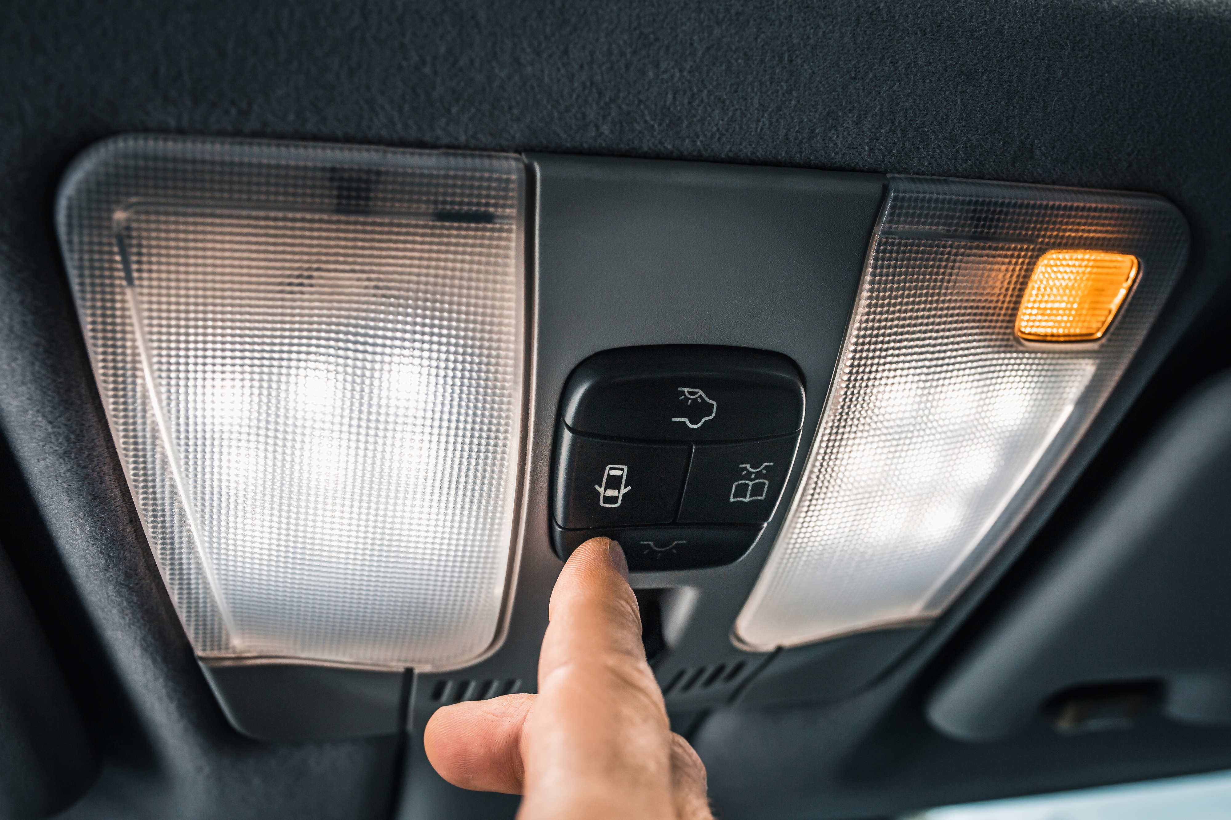 A hand presses a button on a car's interior console, surrounded by overhead lights