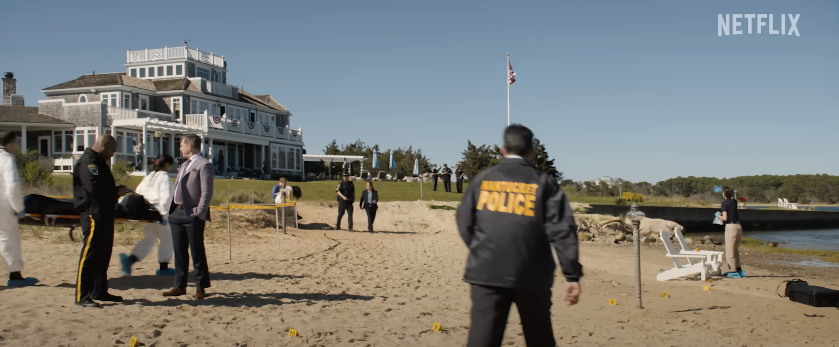 A crime scene near a beach, with investigators and police officers. A large house is in the background. Text in the top right reads "NETFLIX."