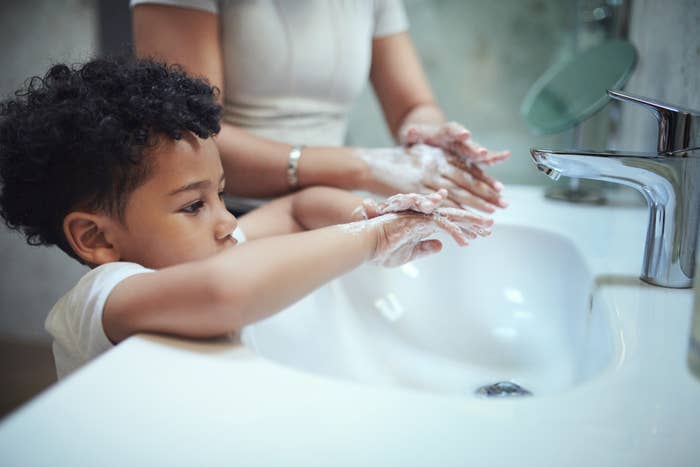 A child with an adult washes hands at a sink, focusing on proper hygiene. Both have soap on their hands