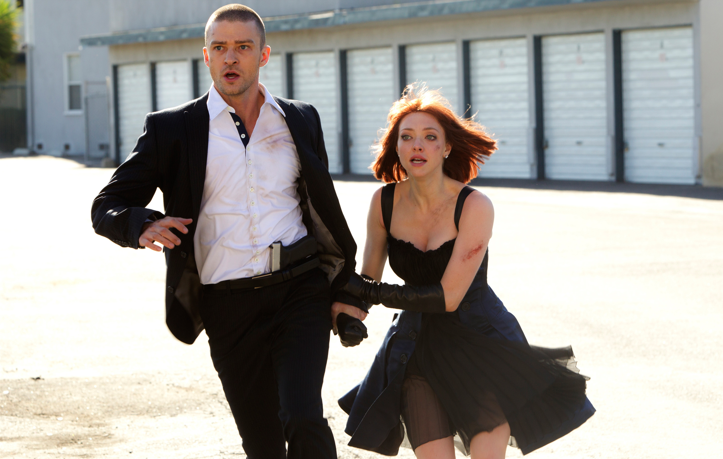 Justin Timberlake in an untucked dress shirt and suit and Amanda Seyfried in a black dress and long gloves run hand-in-hand in a parking lot in a scene from &quot;In Time&quot;