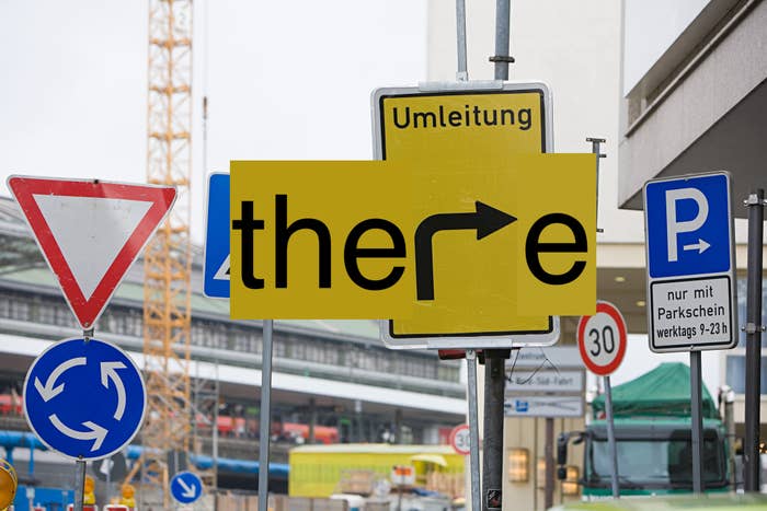 Road traffic signs including detour (Umleitung in German), parking and turn signs form the word &quot;there.&quot; Busy street background with construction crane visible