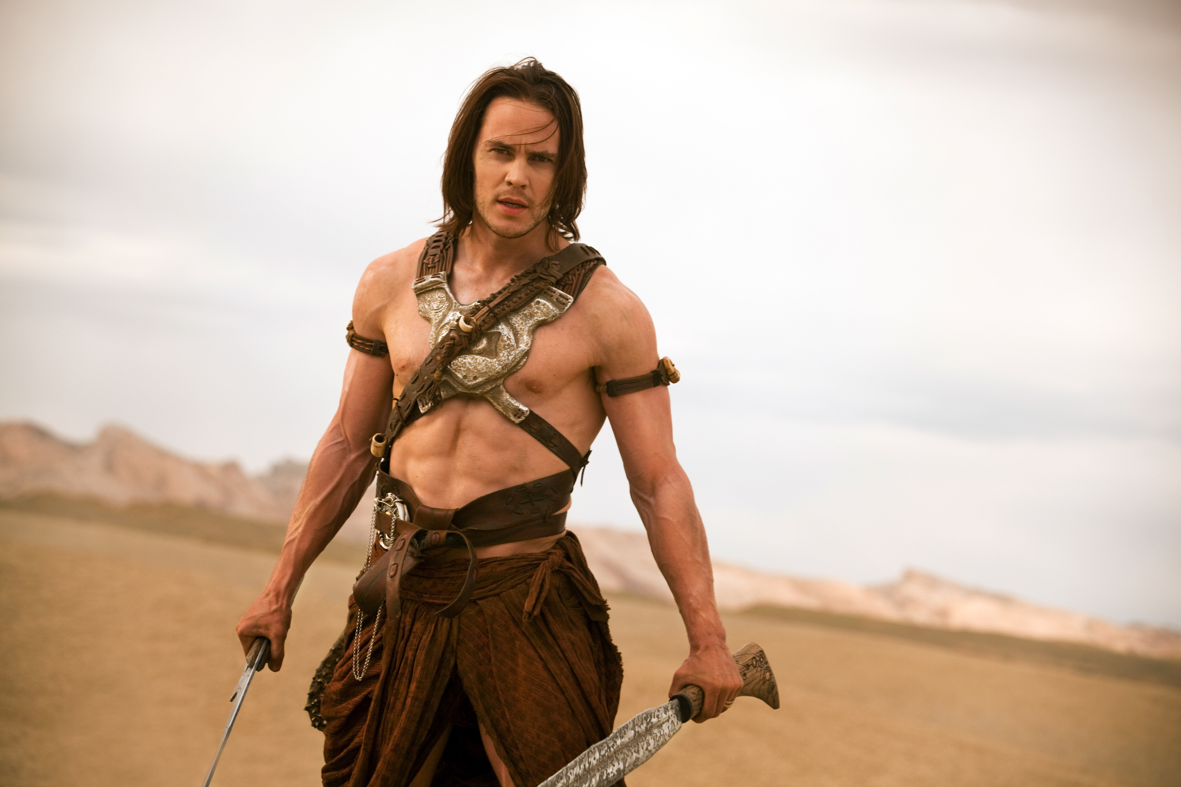 Taylor Kitsch, dressed as John Carter, stands in a desert holding a sword, with armor straps across his chest, in a scene from the movie &quot;John Carter&quot;