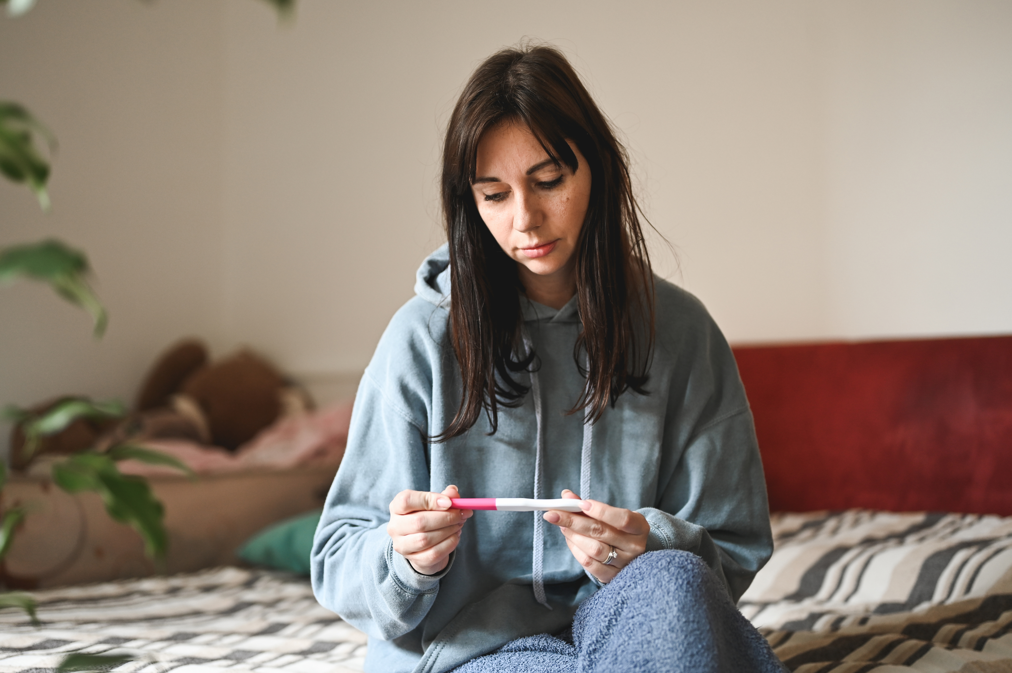 Woman in a hoodie sits on a bed, holding and looking at a pregnancy test