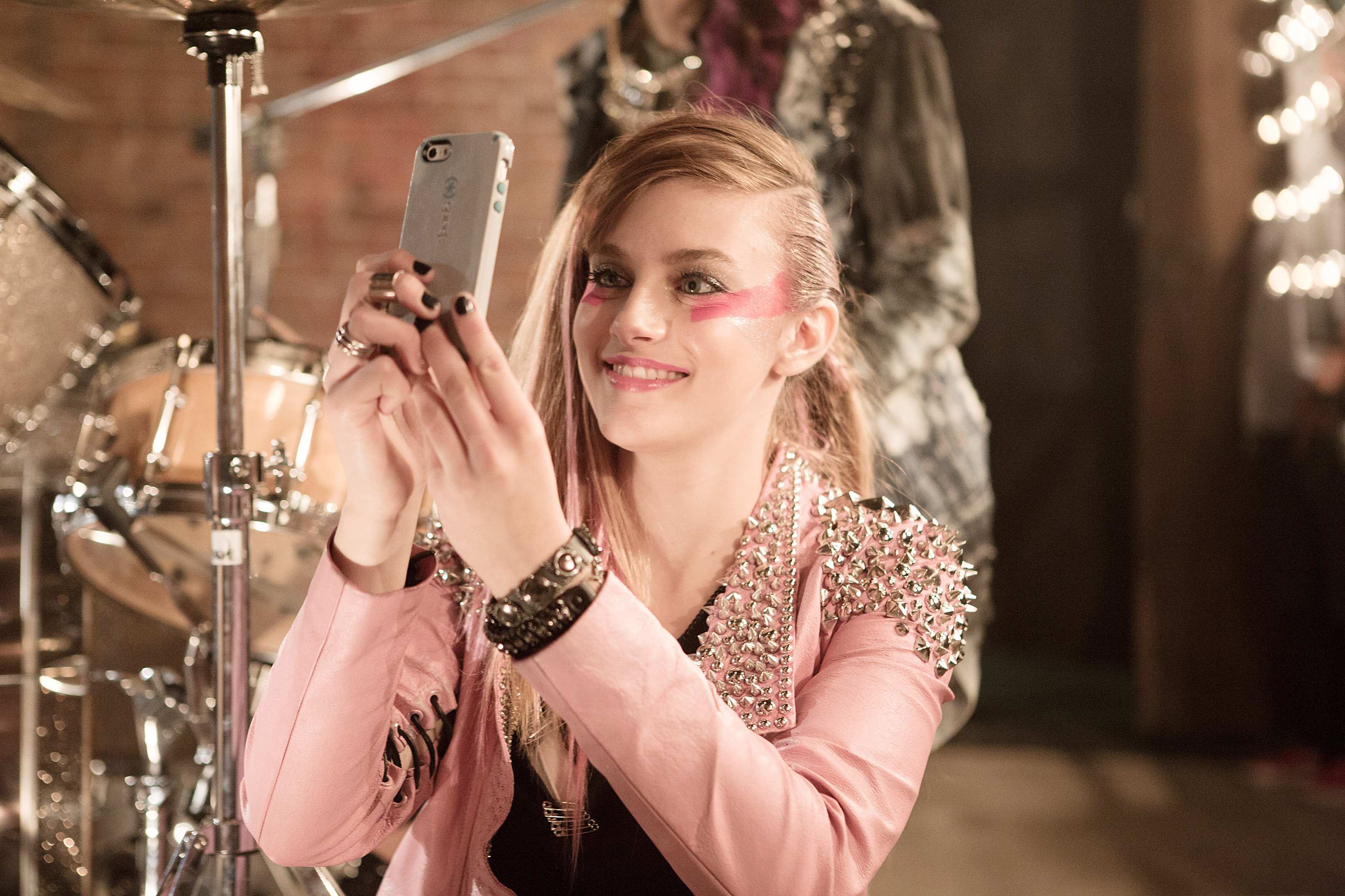Aubrey Peeples in a pink jacket with pink makeup on her face holding a cell phone with drums behind her in a scene from &quot;Jem and the Holograms&quot;