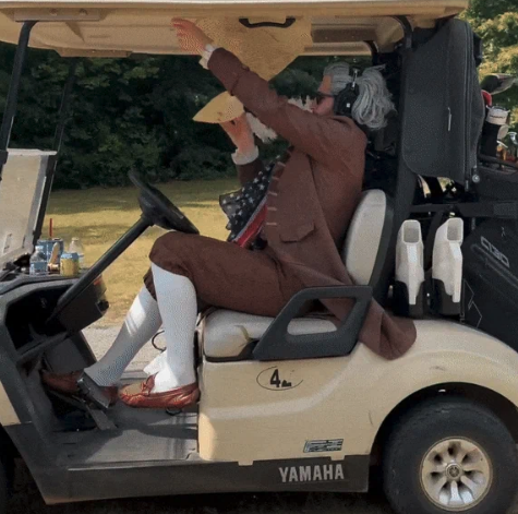 A person dressed in historical attire, including a brown coat, white stockings, and a wig, is sitting in a golf cart and adjusting the roof