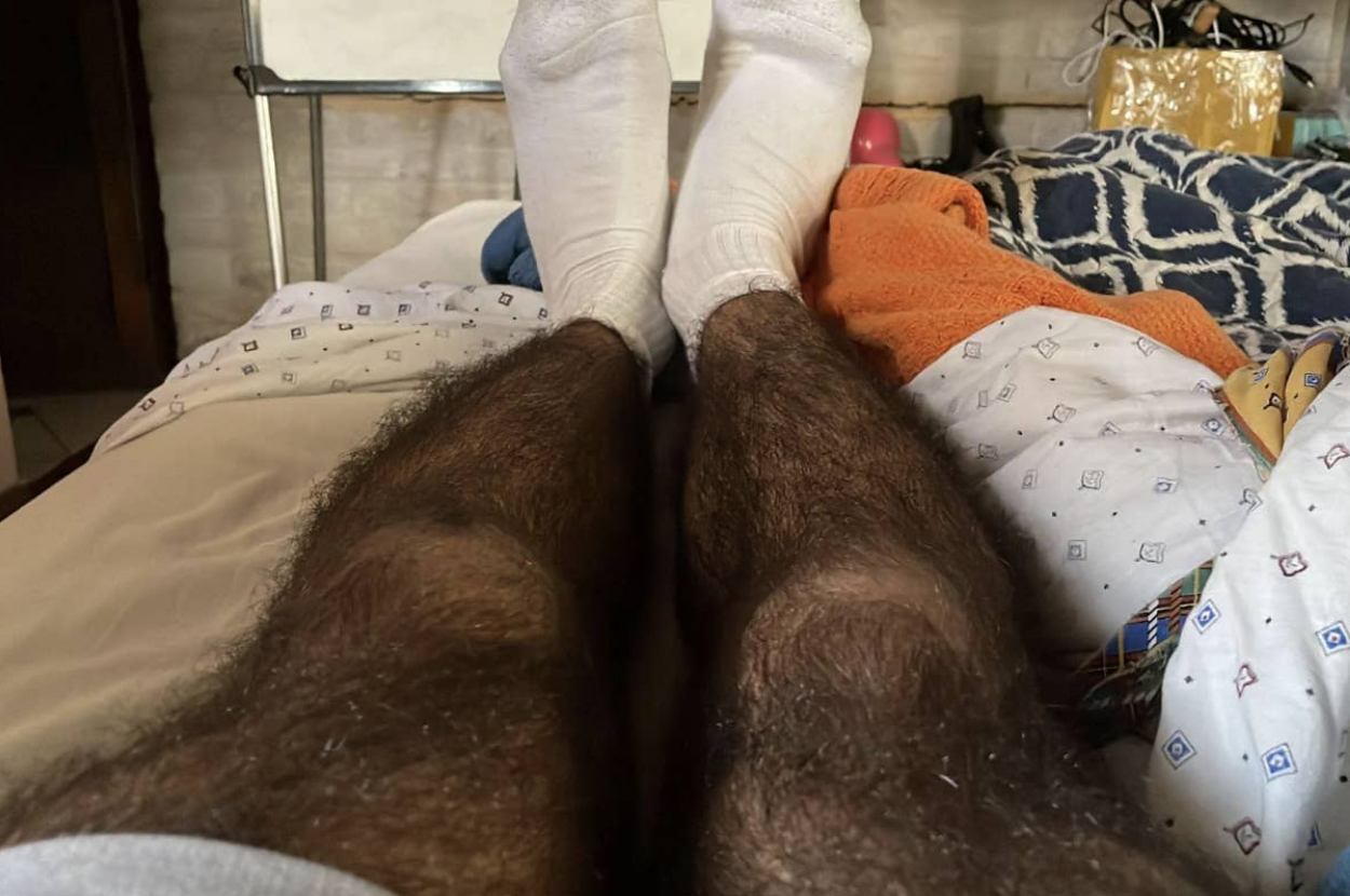 Person lying in bed with socks on, legs visible. Background includes a blanket, pillow, and assorted items