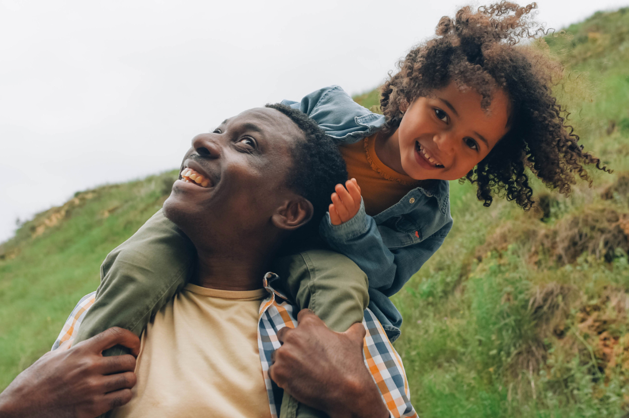 A smiling man carries a happy young child on his shoulders outside in a nature setting. Both are casually dressed in light shirts and jackets