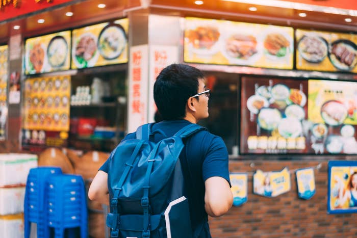 A person with a backpack stands in front of a street food stall featuring various dishes displayed on overhead menus