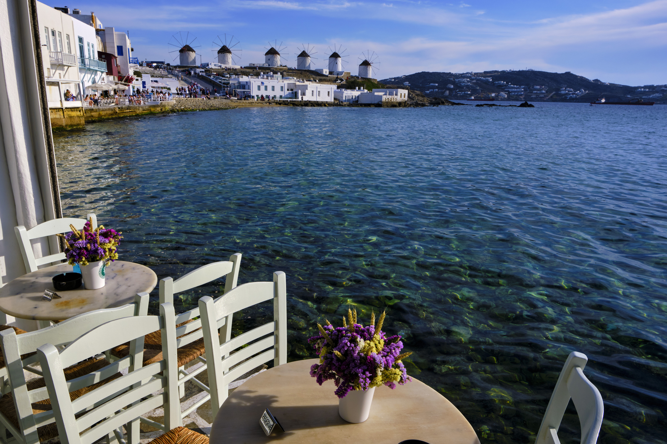 Outdoor seating at a seaside cafe overlooking crystal-clear water and distant traditional windmills in Mykonos, Greece, with tables adorned with purple flowers